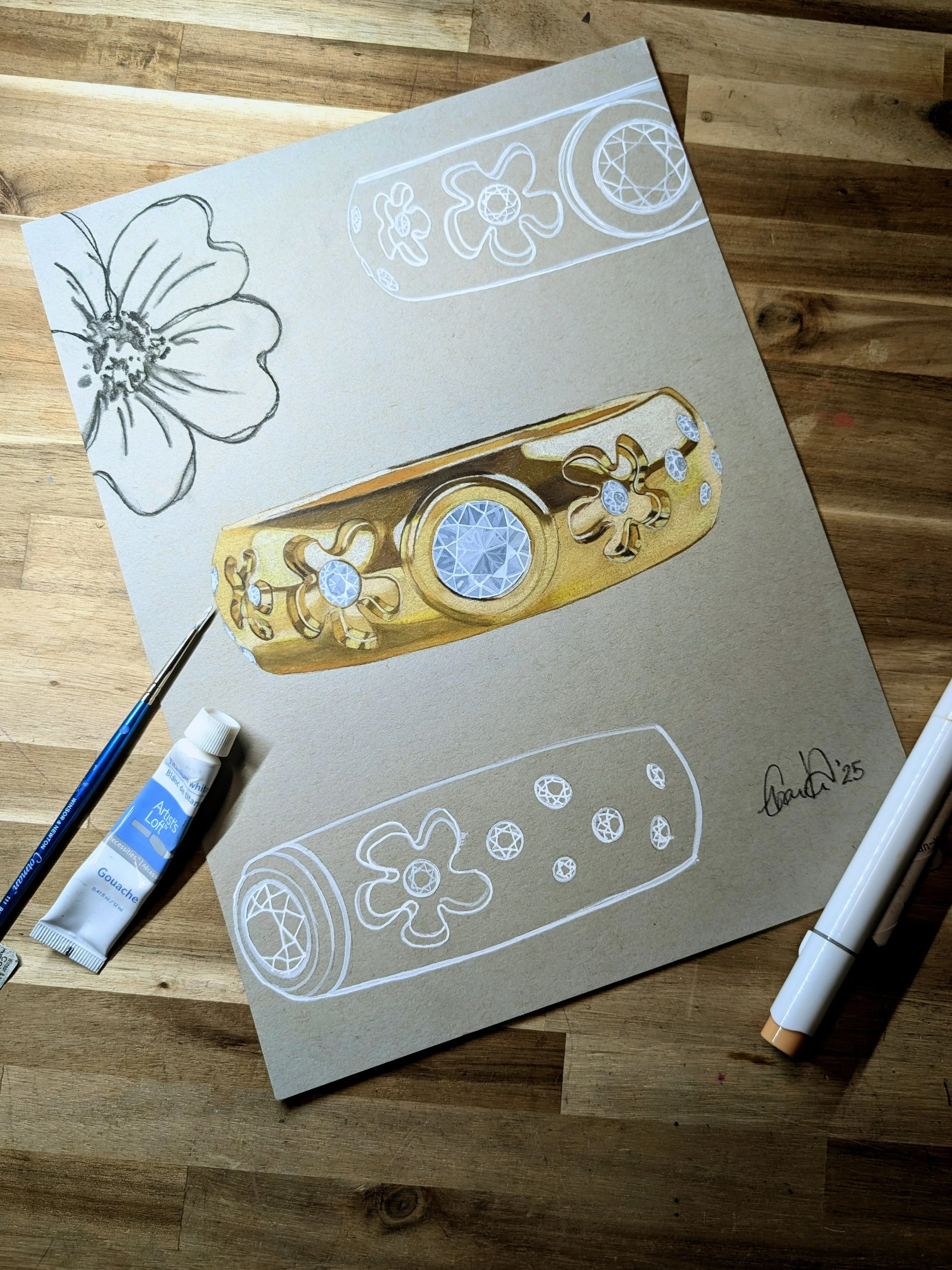 Artwork of a gold ring with gemstone and floral engravings, displayed on textured paper with sketches of the ring and a flower, surrounded by art supplies on a wooden surface.