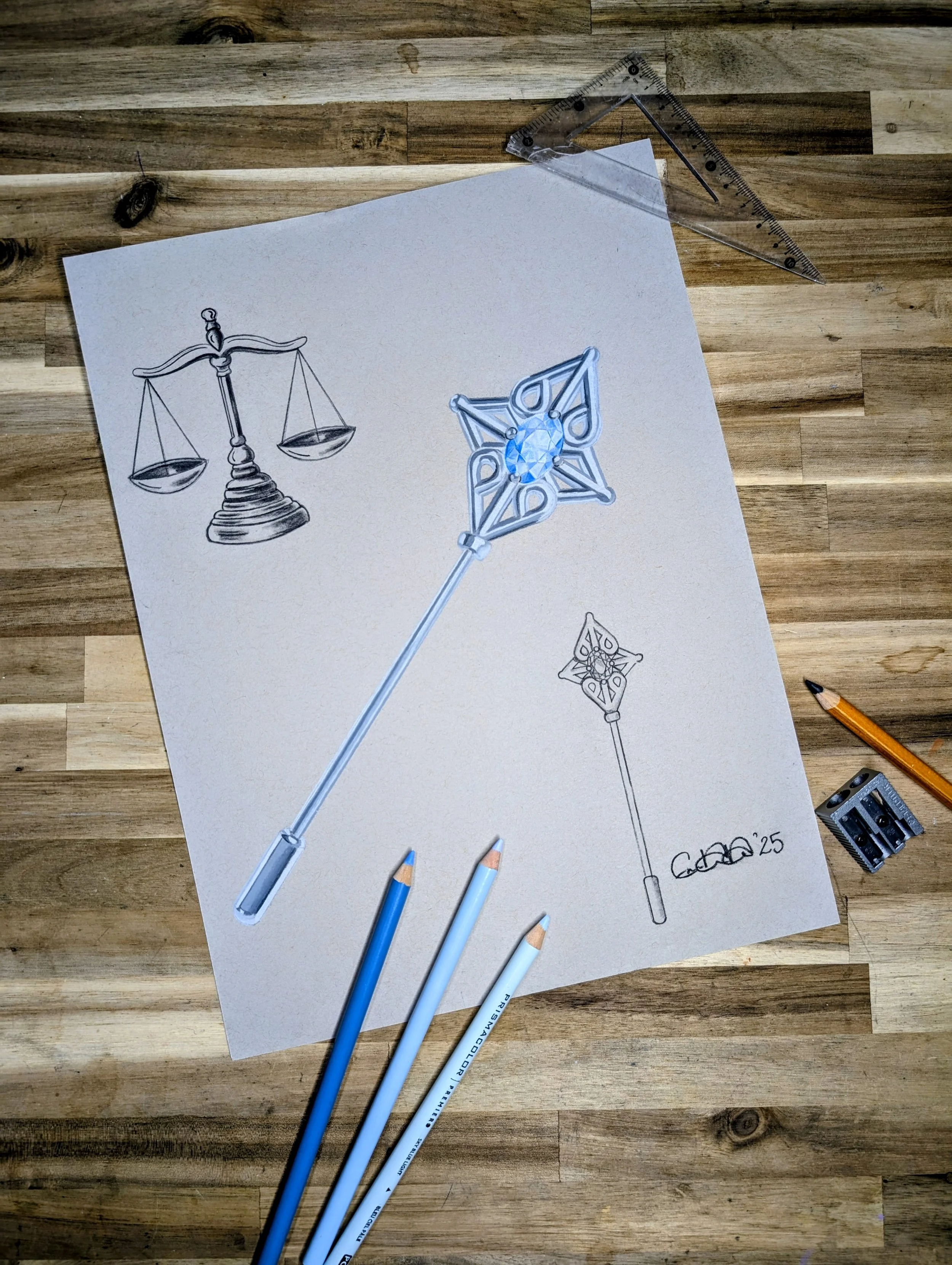 A sheet of paper with sketches of a lapel pin, with the scales of libertysurrounded by colored pencils, a pencil, a precision knife, a square ruler, and a triangle ruler on a wooden surface.