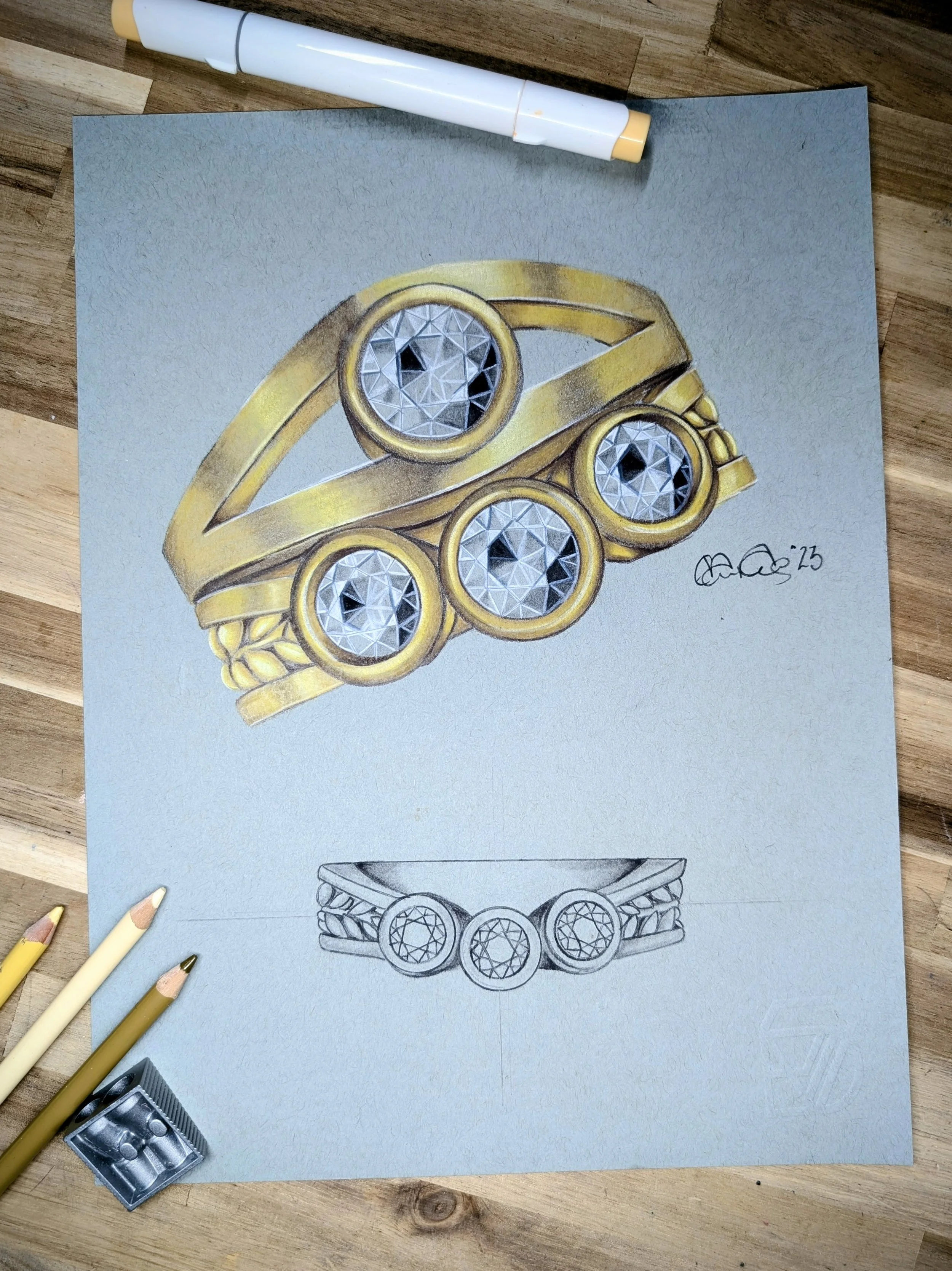 A detailed drawing of a gold ring with multiple round diamonds, some with intricate faceting, and a pencil sketch of the same ring design beneath it, on a metal surface with drawing pencils and a sharpener nearby.