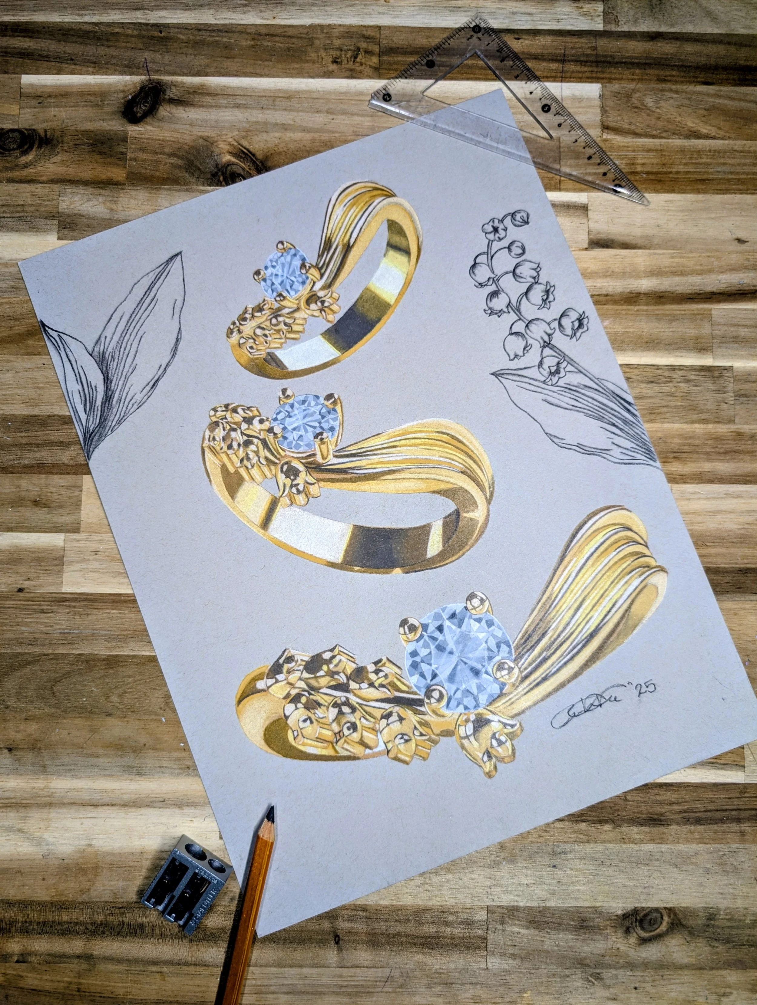 A detailed jewelry design illustration featuring gold rings with diamonds and floral accents, on light gray paper with sketches of leaves and flowers, placed on a wooden surface with drafting tools nearby.