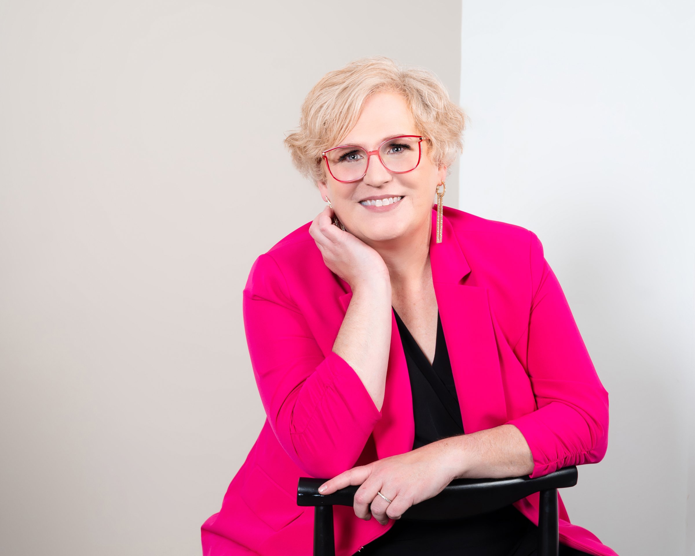Person in a bright pink blazer and glasses smiling and resting their head on their hand, seated against a neutral background.