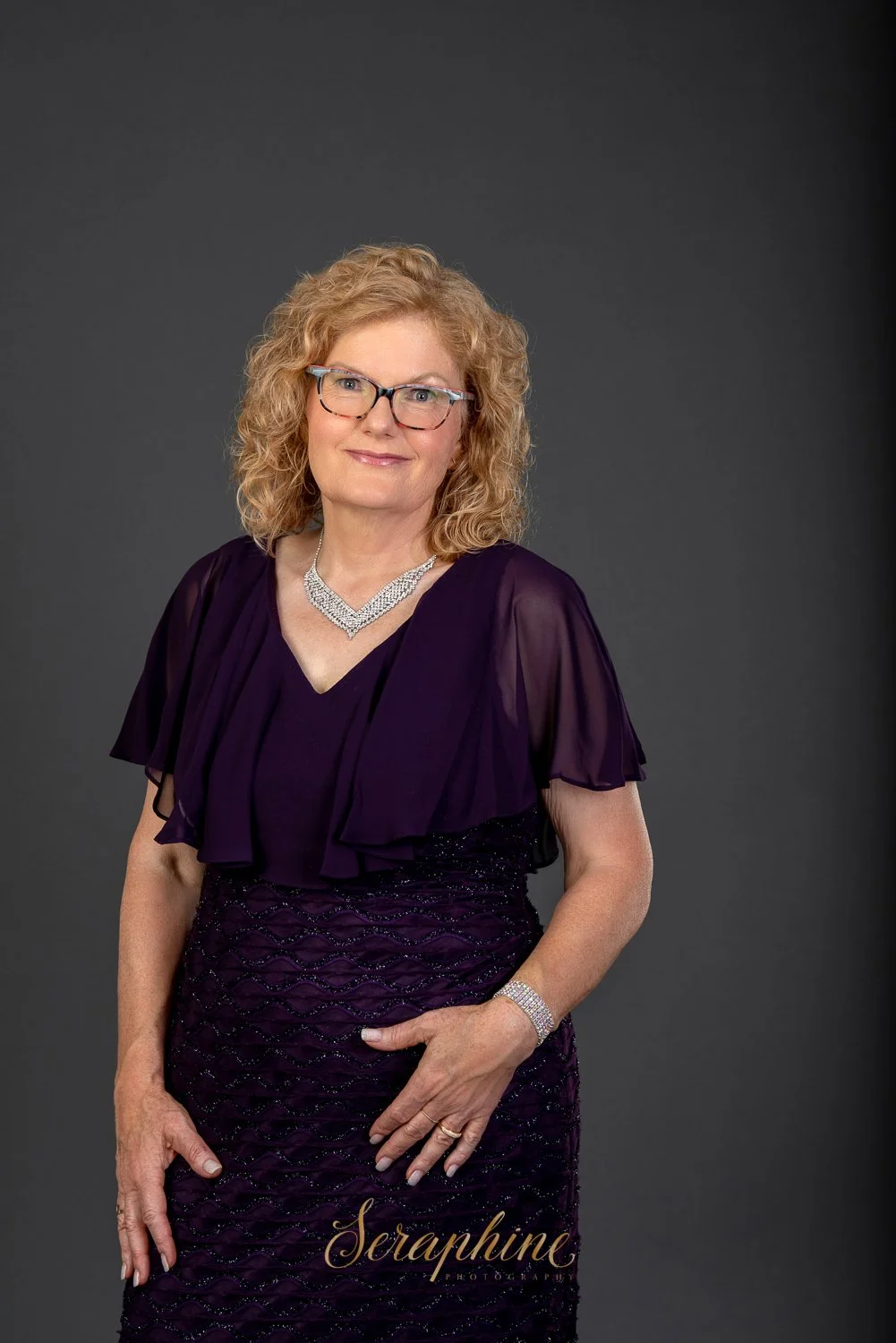 Woman with curly hair wearing glasses and a purple dress, standing against a gray background.