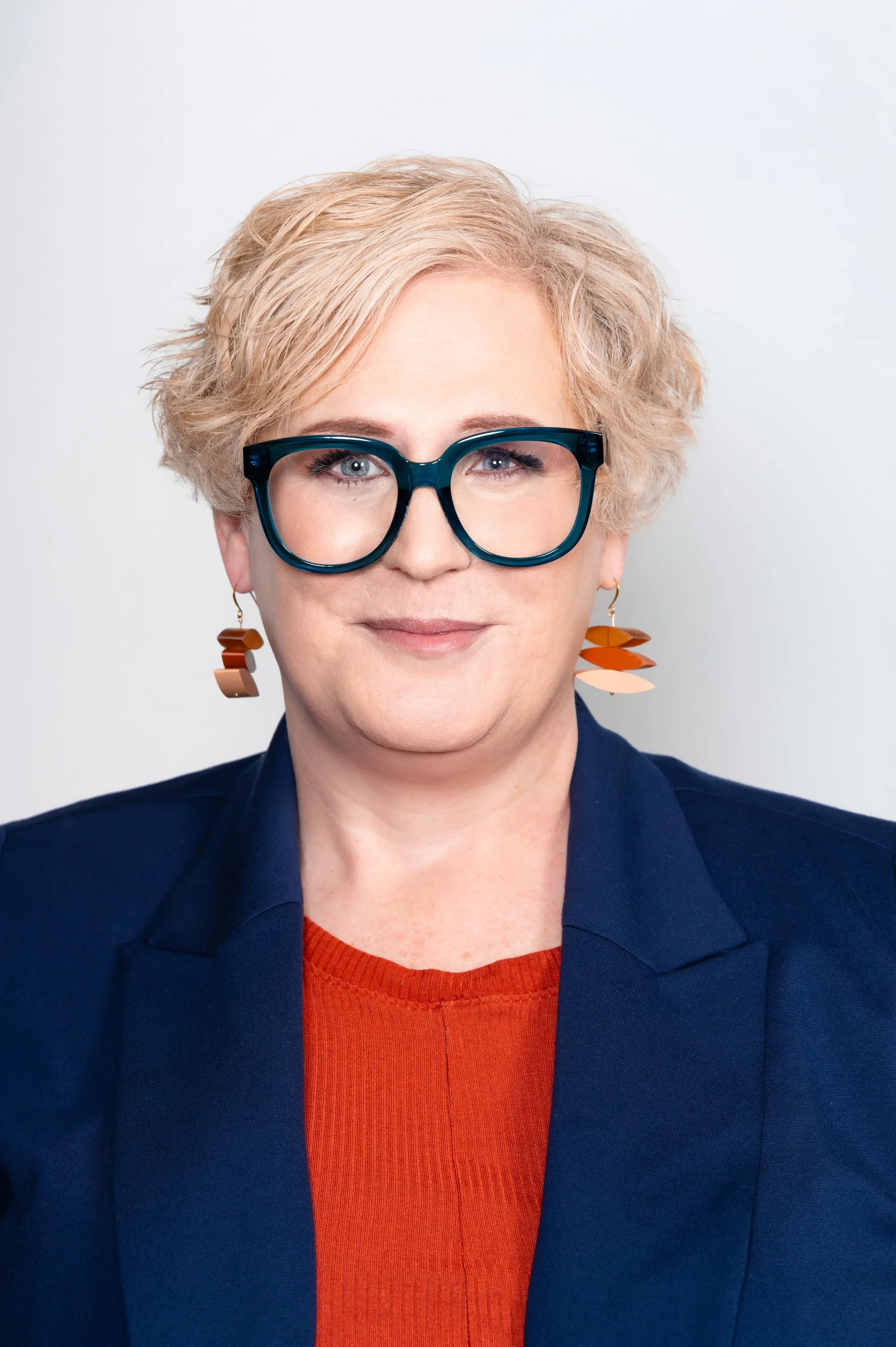 Person with short blonde hair, wearing large blue glasses and geometric orange earrings, dressed in a navy blazer and orange top.