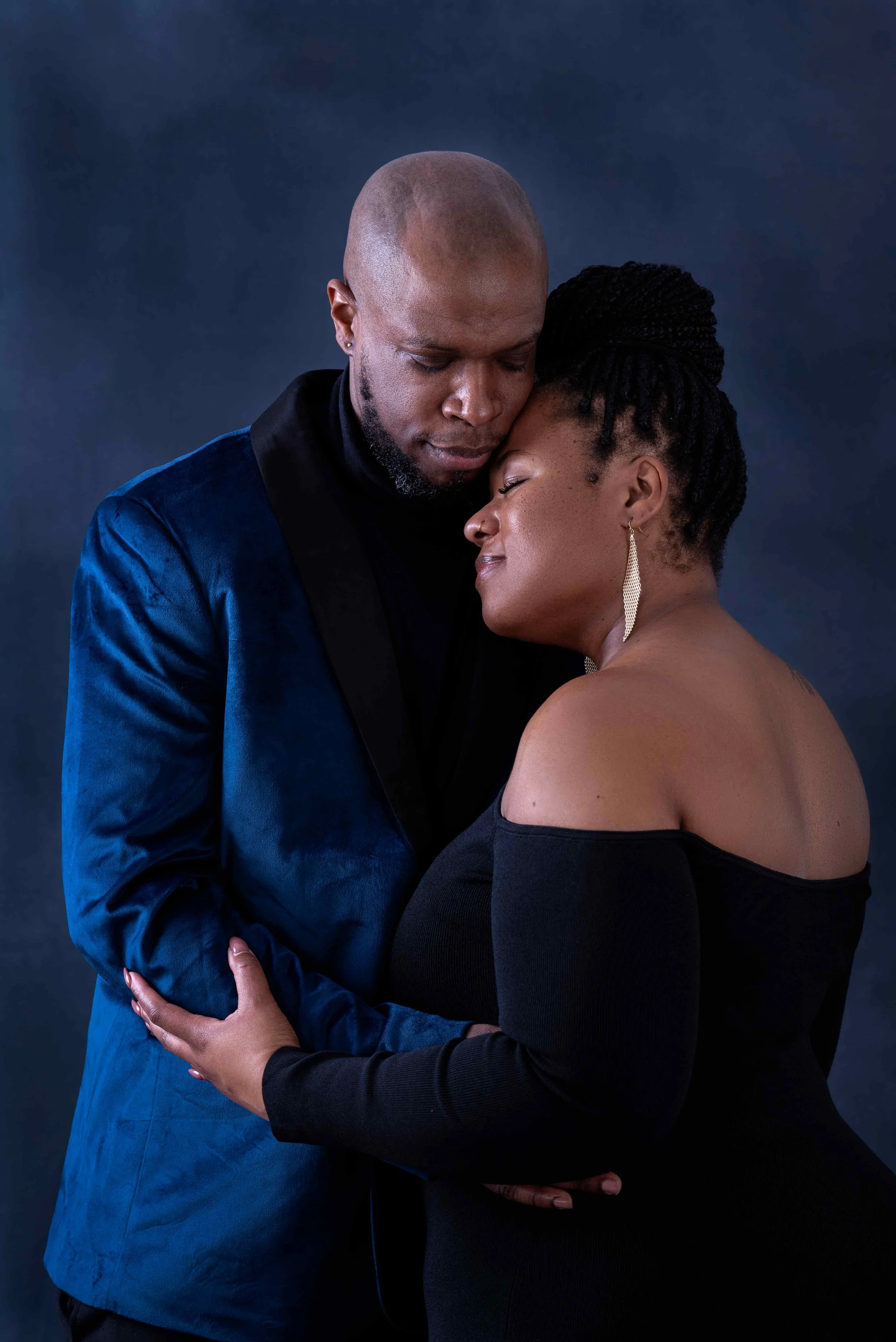 A couple embracing intimately in front of a dark backdrop. The man is wearing a blue velvet jacket, and the woman is in a black off-shoulder dress, showing affection and closeness.
