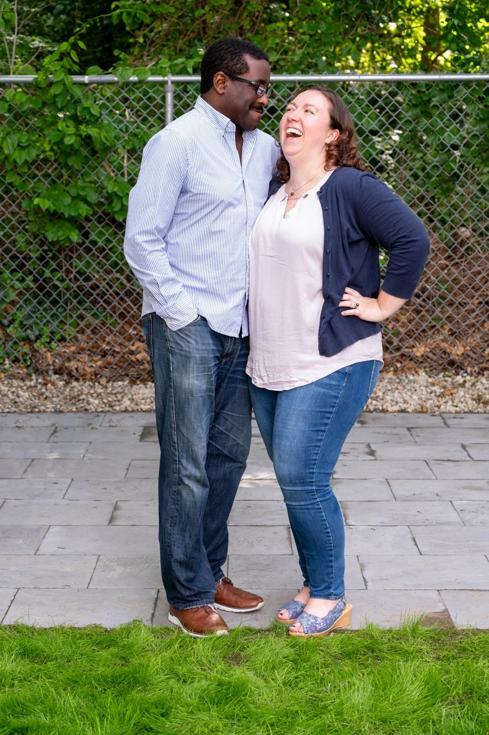 A man and woman laughing together outdoors in a garden, standing on a stone patio with greenery and a chain-link fence in the background. They are wearing casual, coordinated outfits with jeans and button-down shirts.