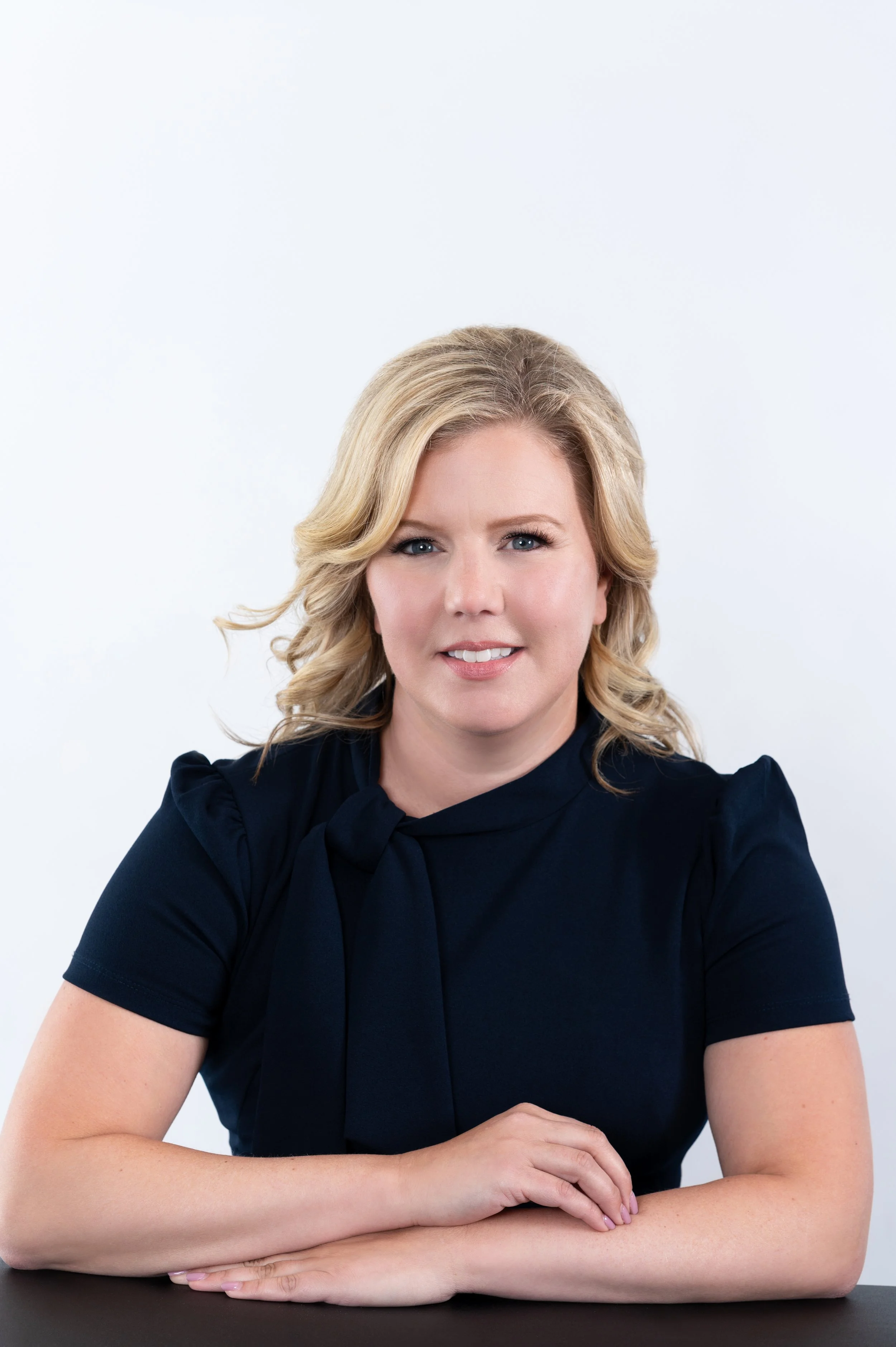 Portrait of a person with blond hair wearing a dark navy blouse, sitting with hands crossed on a table against a white background.