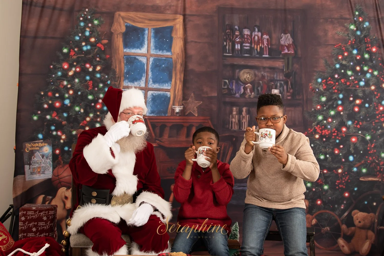 Santa Claus and two children drinking from mugs in a cozy, festive Christmas setting with a decorated tree and holiday backdrop.