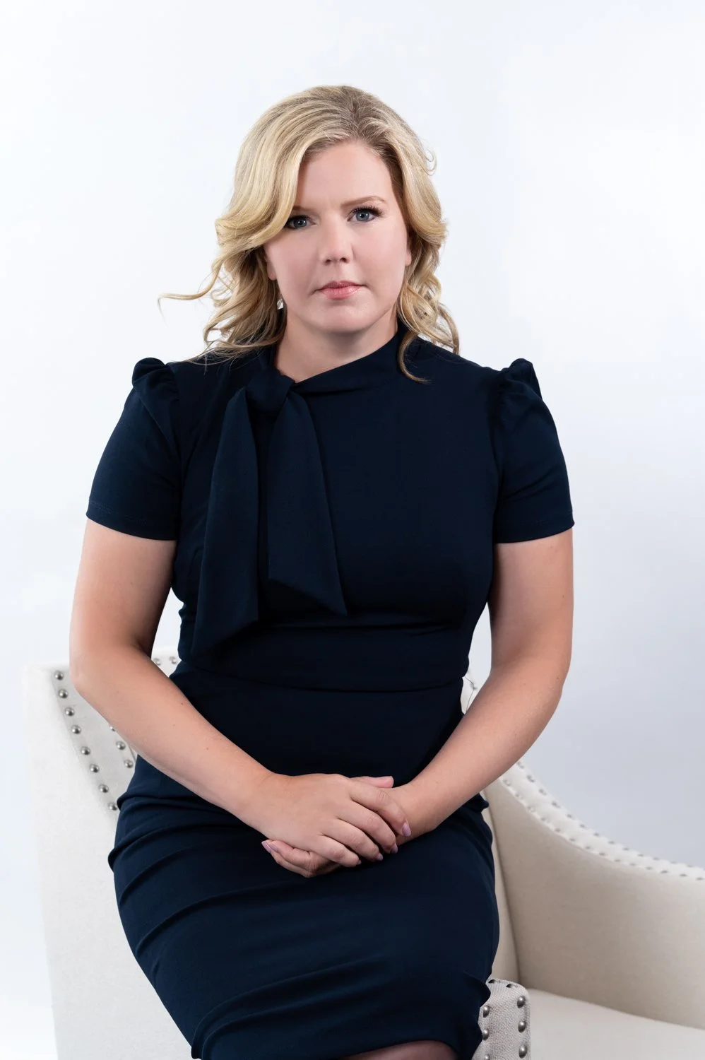 Woman sitting on a chair wearing a dark dress, with arms folded on her lap.