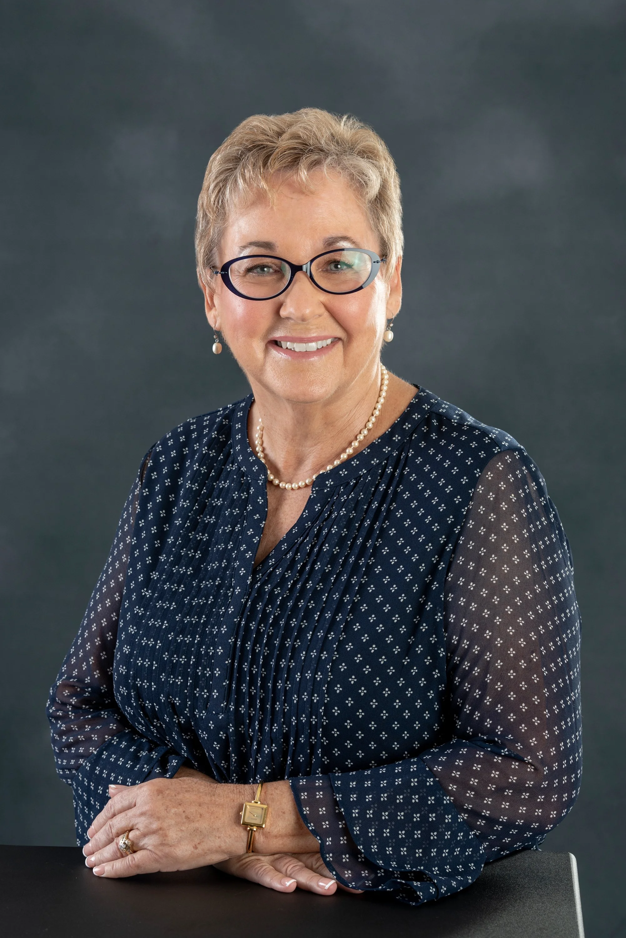 Smiling woman with short hair and glasses wearing a blue patterned blouse, pearl necklace, and gold watch, sitting against a dark background.