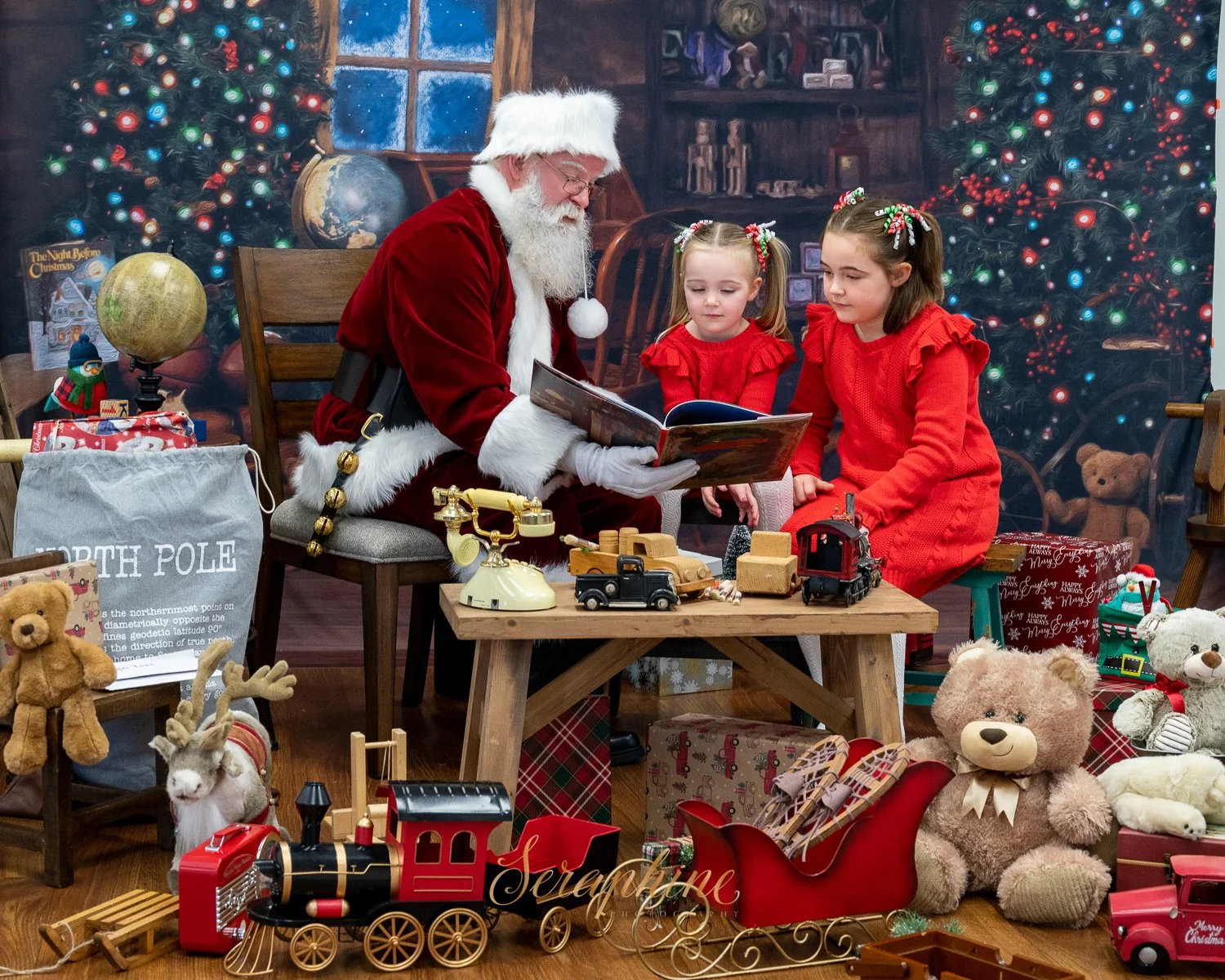 Santa Claus reading a book to two children in red outfits, surrounded by Christmas decorations, toys, a sack labeled 'North Pole,' and a decorated tree.