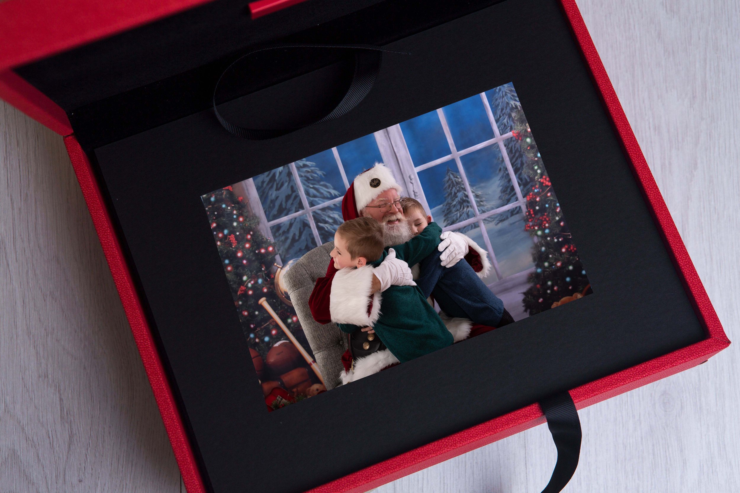 Photo of Santa Claus hugging two children inside a red gift box, Christmas setting with a decorated tree and snowy window.