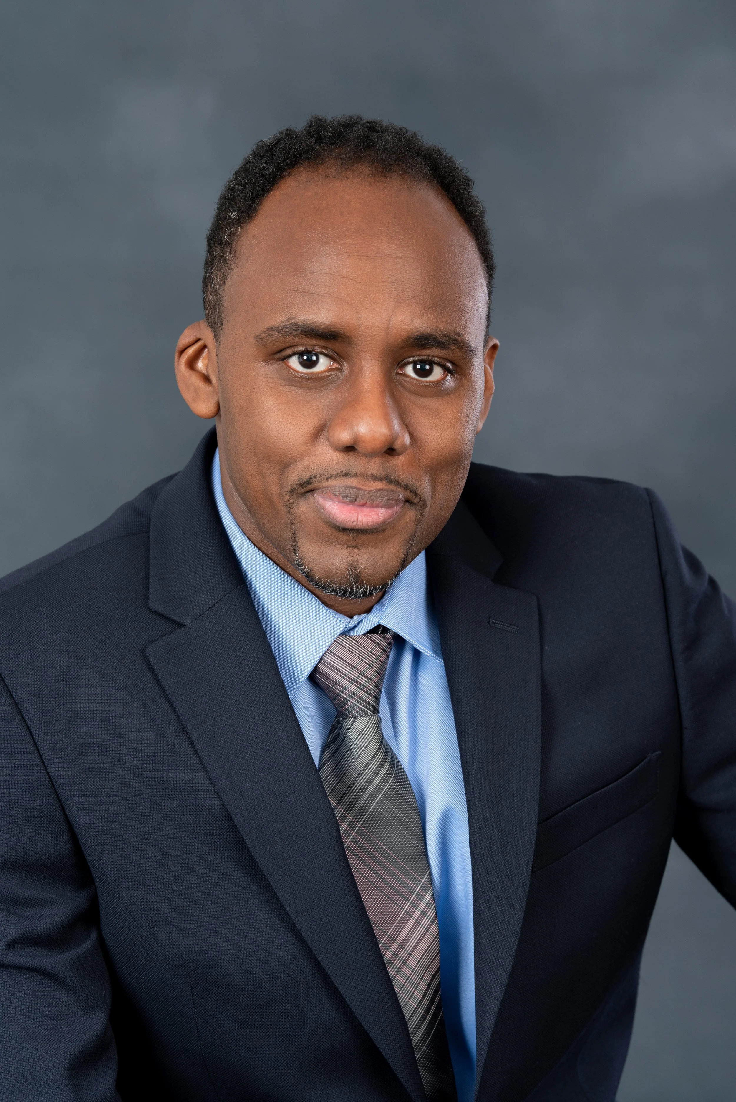 Portrait of a man in a suit with a blue shirt and gray tie, against a gray background.