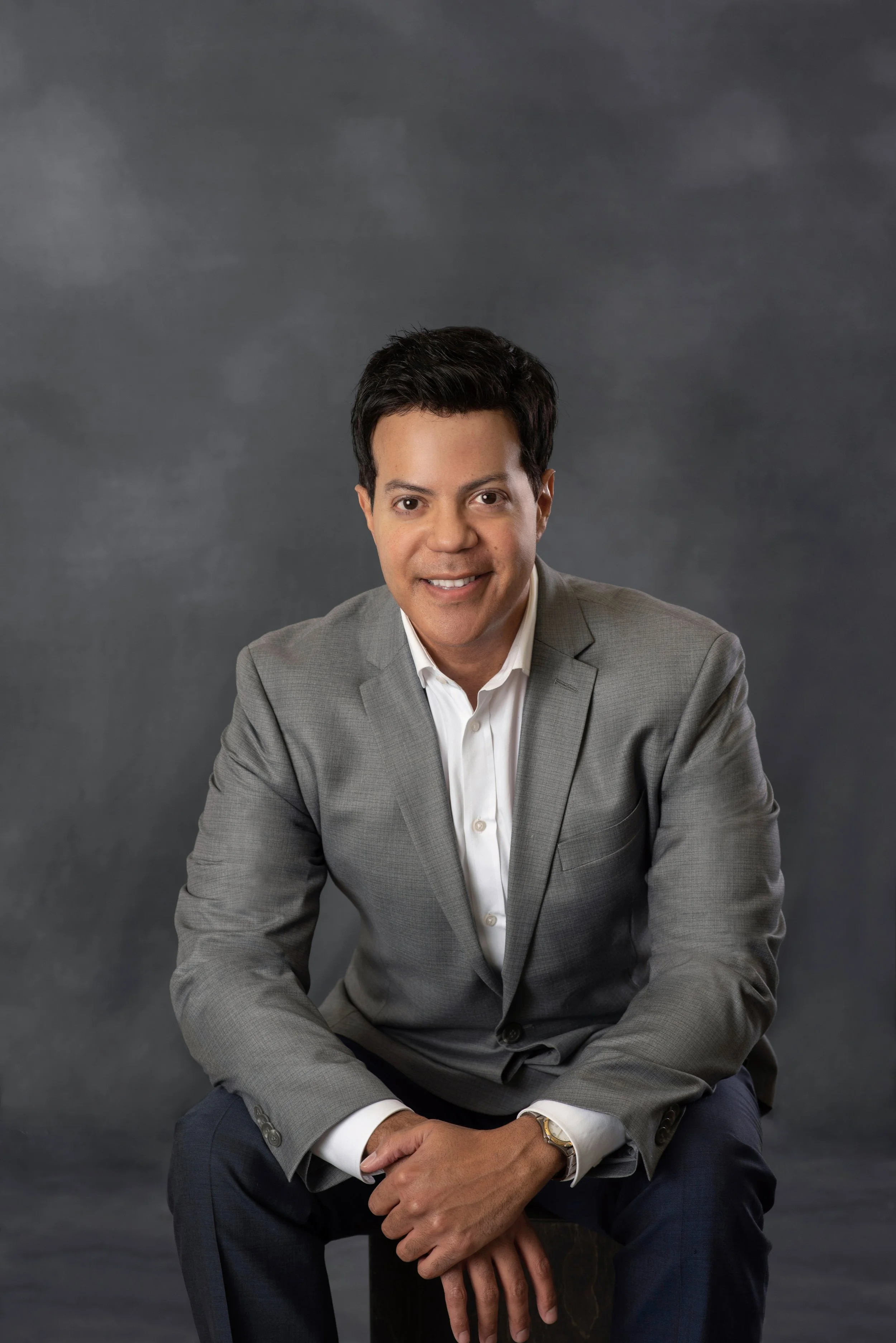Man in a gray suit sitting and smiling against a gray background.