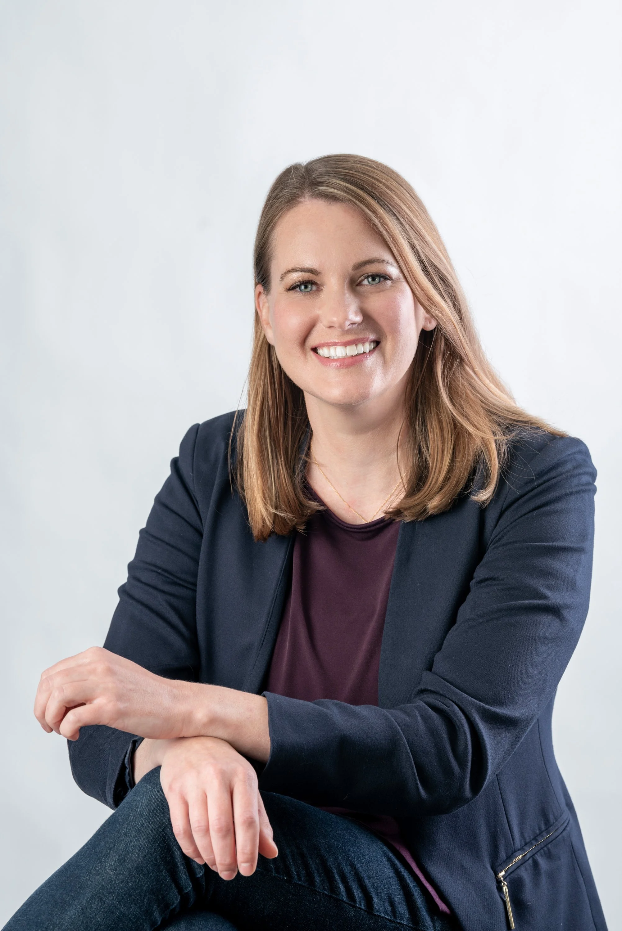 Smiling person with medium-length hair wearing a dark blazer and a maroon shirt against a light background.