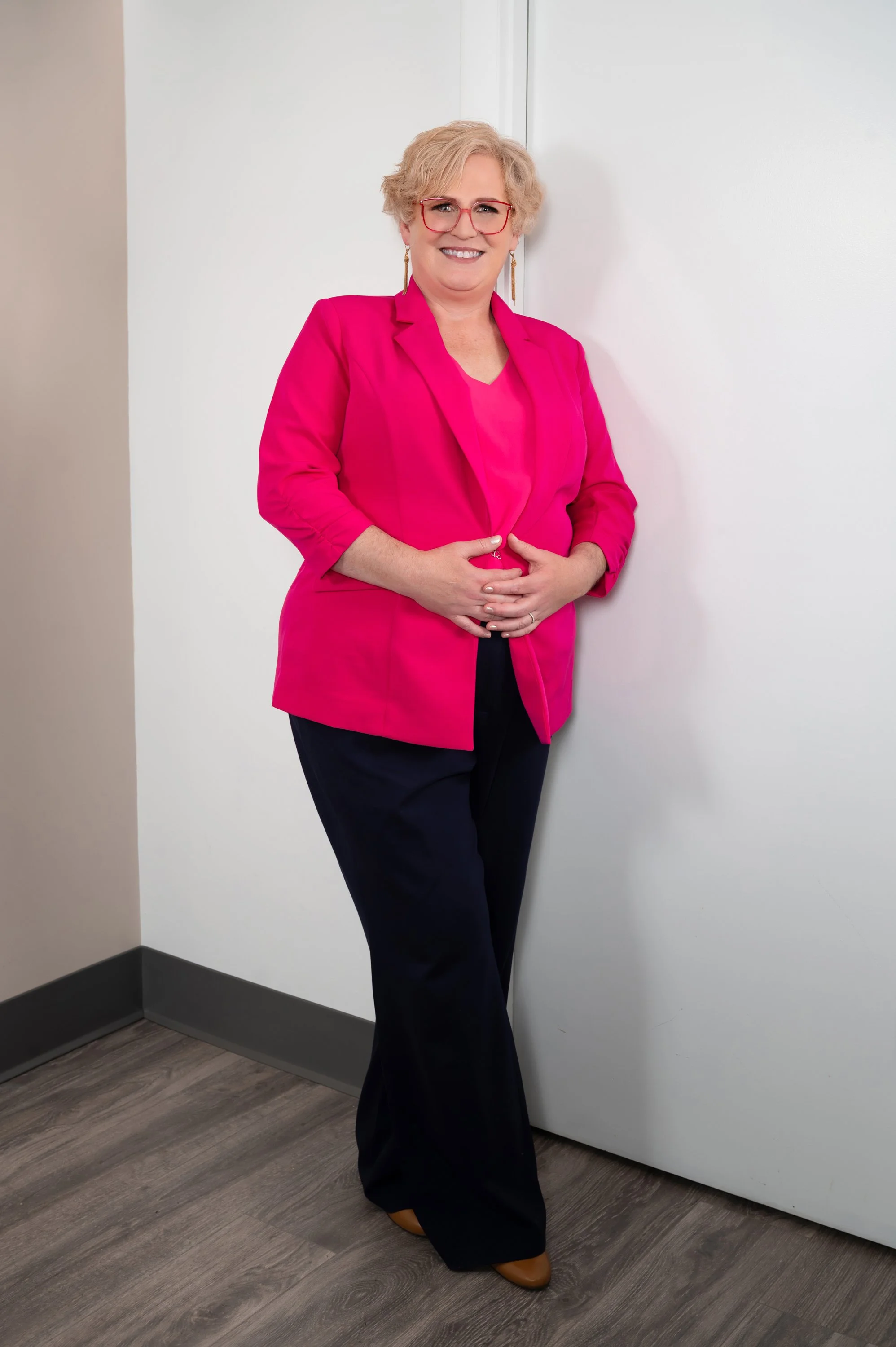 Person with short blond hair wearing a bright pink blazer, navy pants, and glasses, standing in a room with hardwood flooring.
