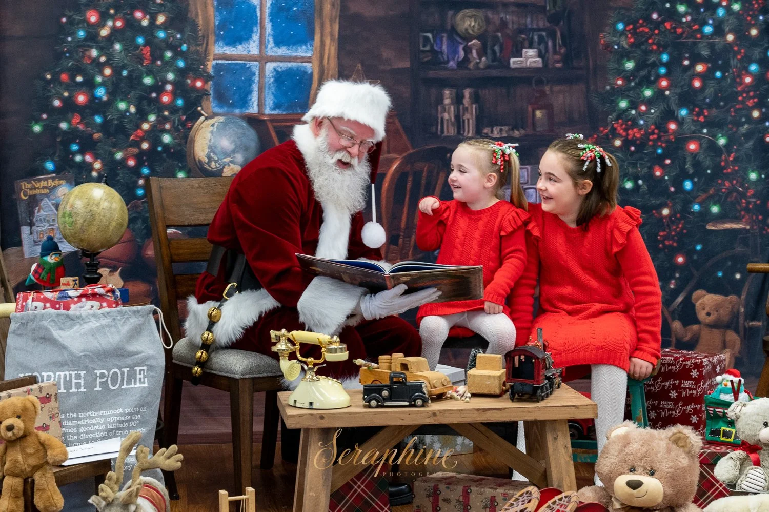 Santa Claus reading a book to two children in red outfits, surrounded by Christmas decorations and toys, with a decorated Christmas tree in the background.