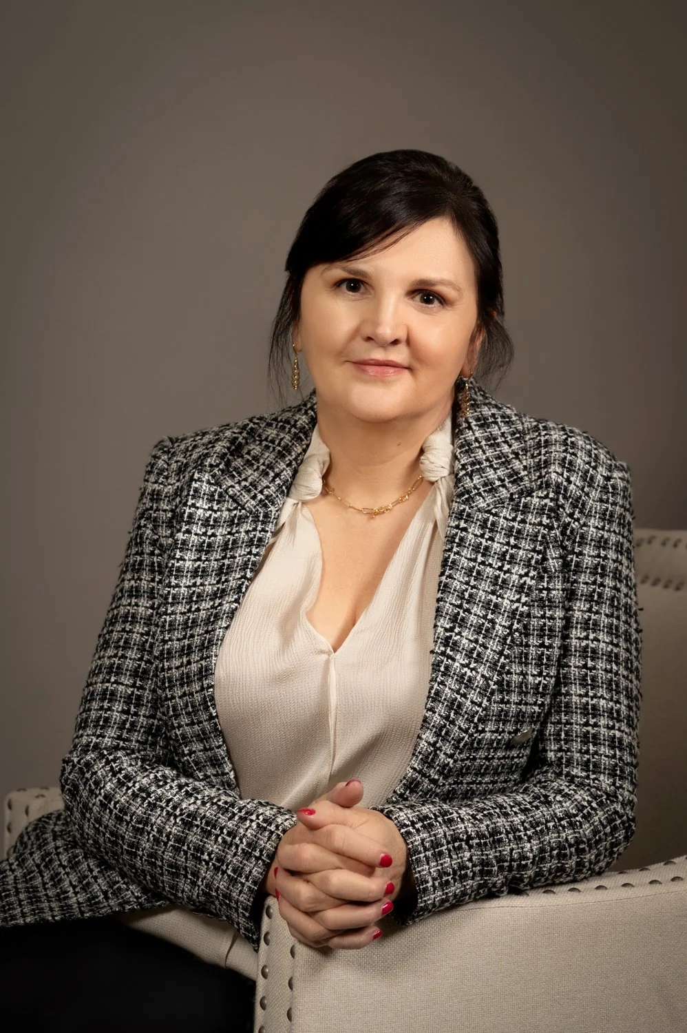 Professional woman sitting, wearing a tweed jacket and cream blouse, with hands folded.