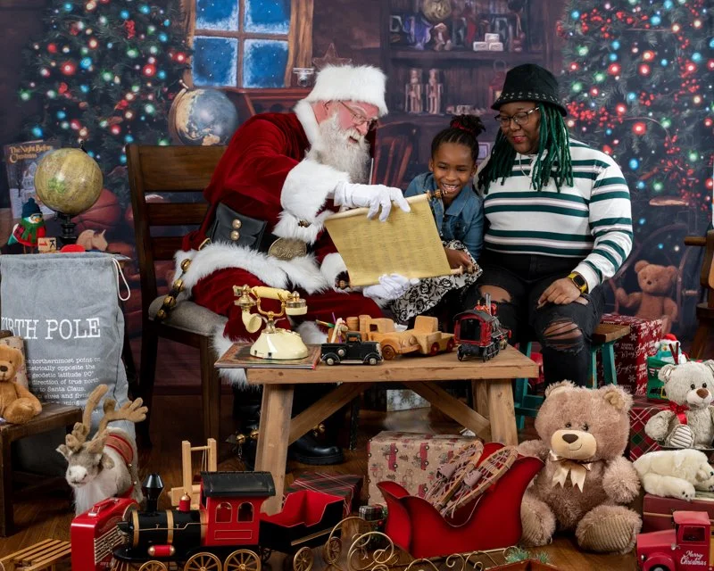 Santa Claus reading a list to a smiling child and adult in a festive room with Christmas decorations, toys, and a Christmas tree in the background.