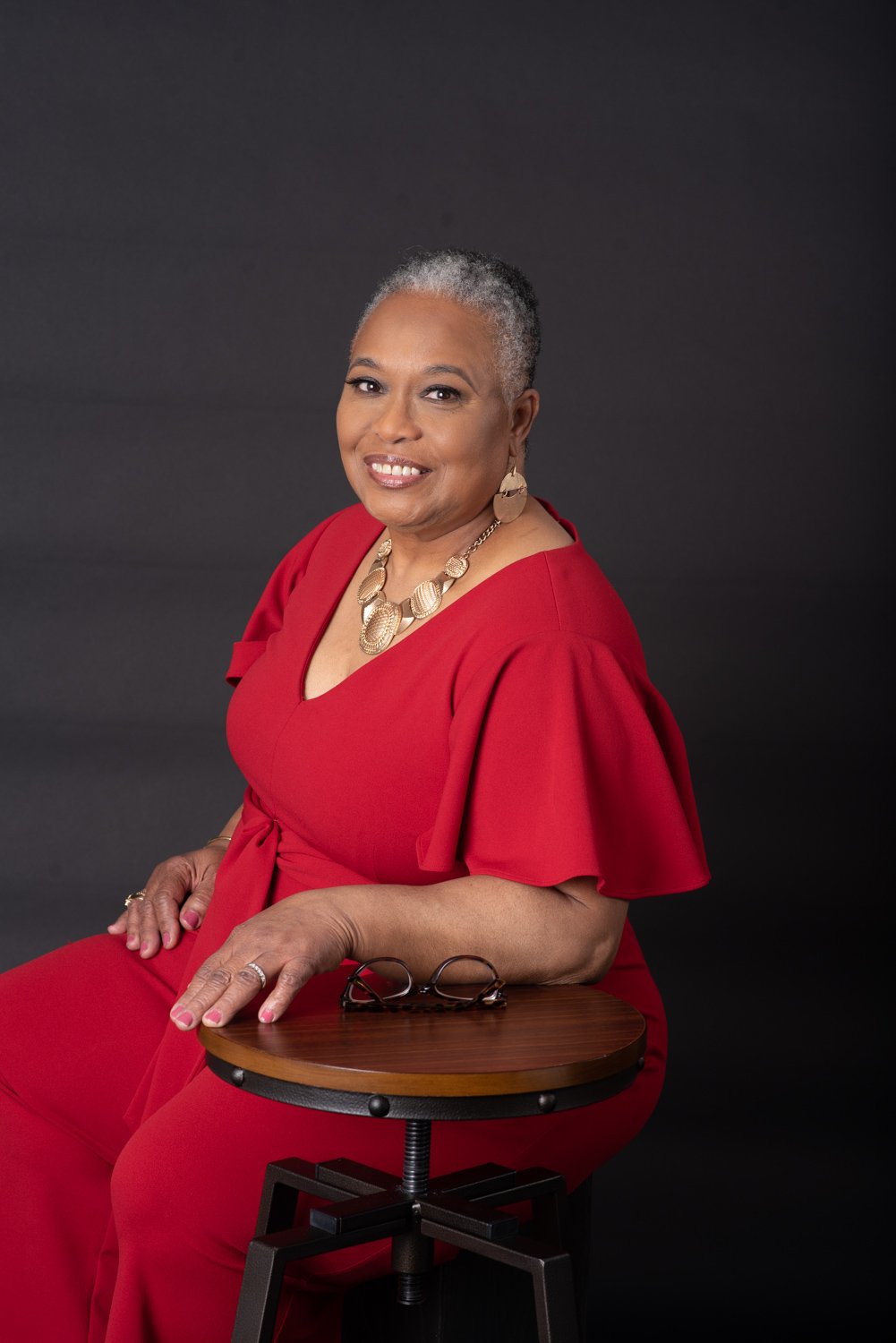 Smiling woman in red dress with jewelry, sitting on a stool against a dark background.