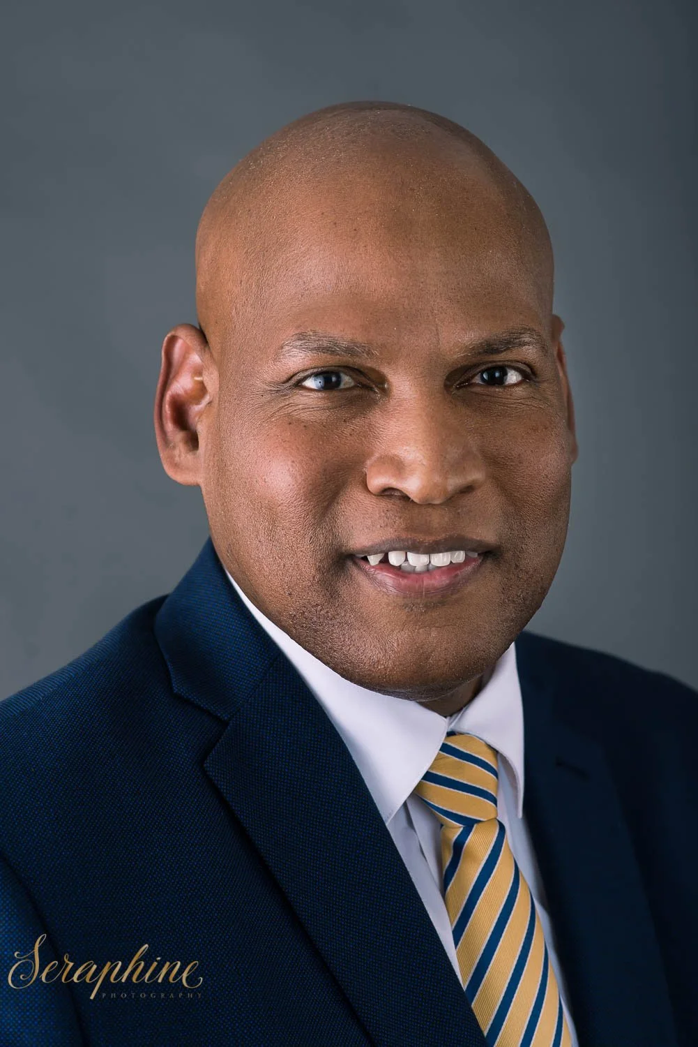 Bald man in a suit and tie smiling for a portrait with a gray background.