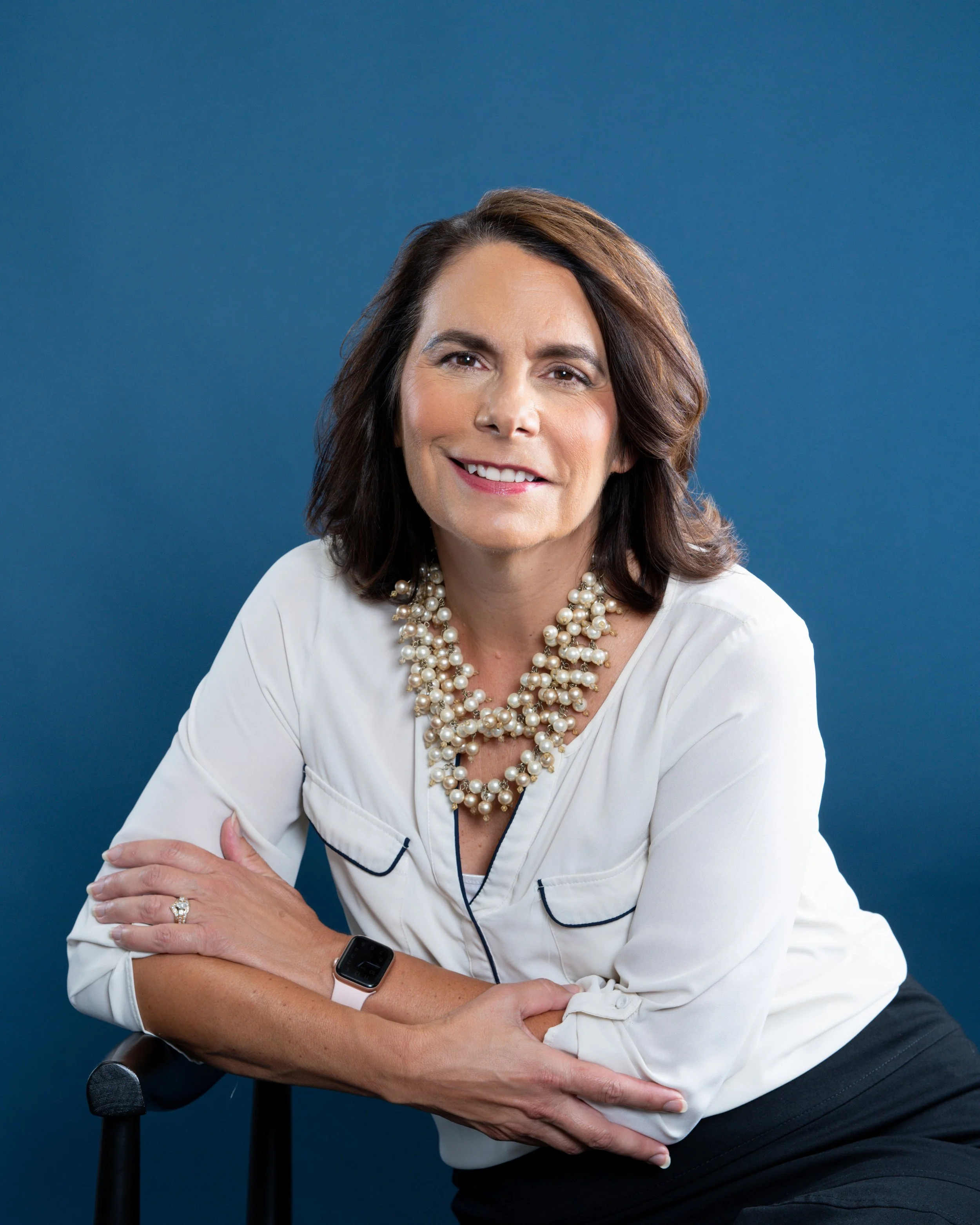A woman with shoulder-length brown hair wearing a white blouse and a layered pearl necklace. She is seated against a blue background, crossing her arms and wearing a smart watch on her left wrist. She is smiling softly.