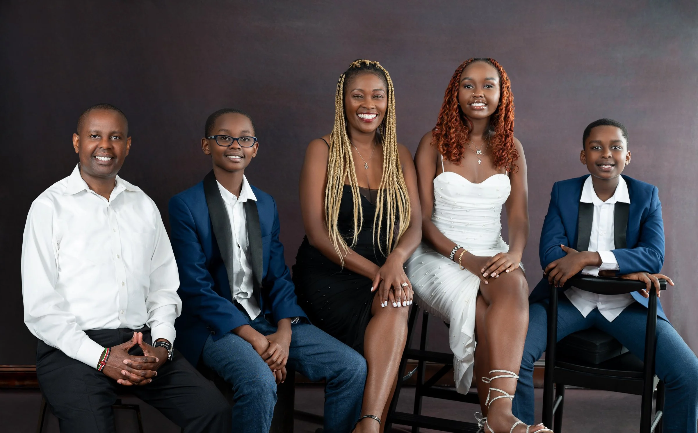 A family of five posing for a portrait in formal attire, with two men in white shirts and black suits, and three women in dresses. The background is dark.