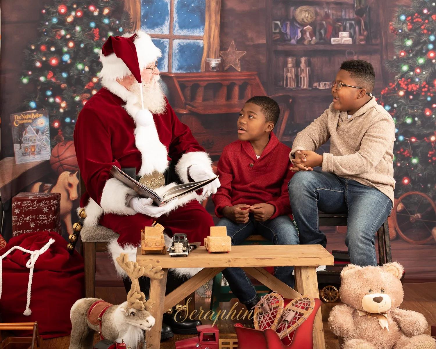 Santa Claus and two children sitting indoors, surrounded by Christmas decorations, with a toy reindeer and teddy bear in the foreground.