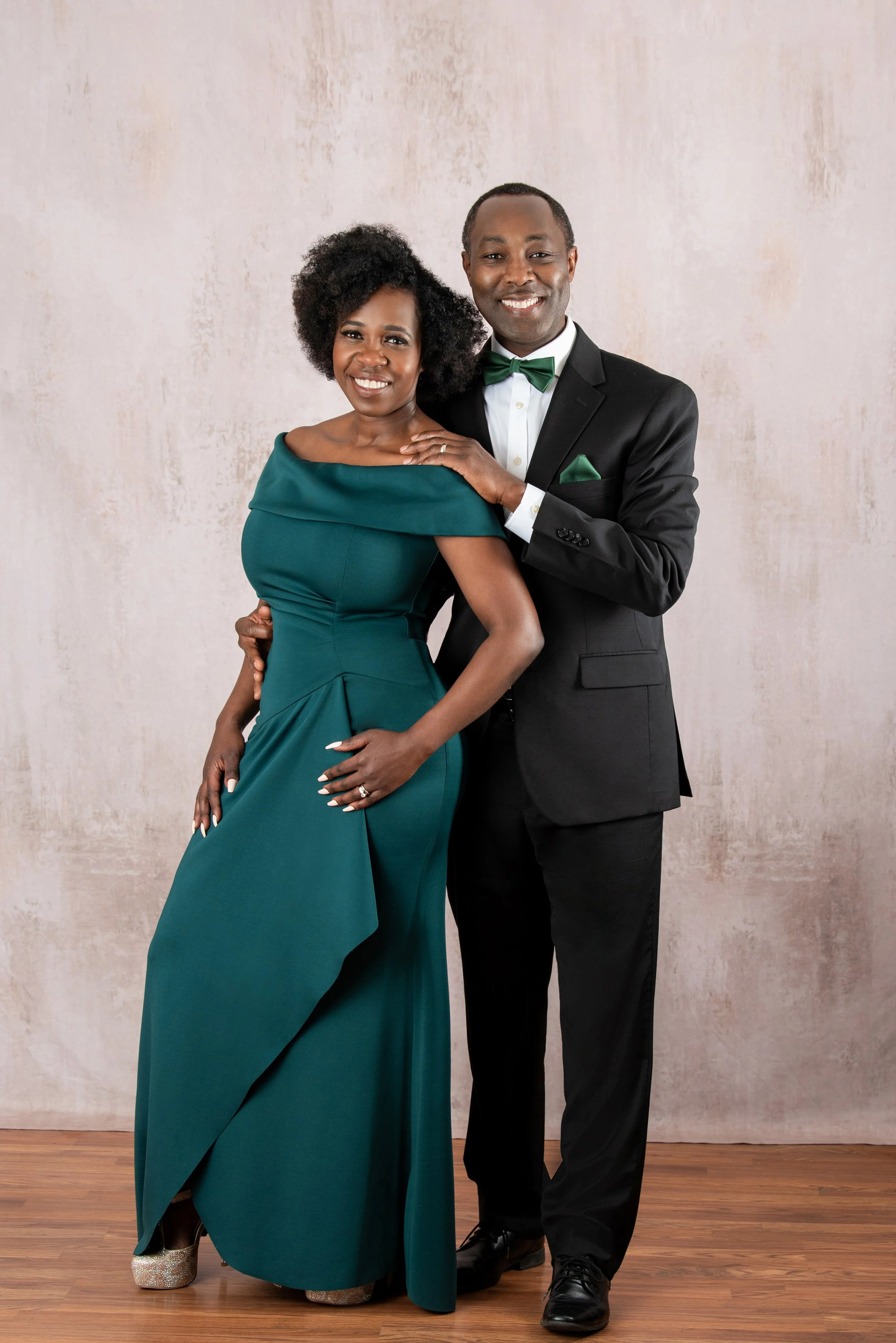 Smiling couple dressed formally, woman in a teal evening gown, man in a black suit with teal accents, posing for a photo against a neutral background.