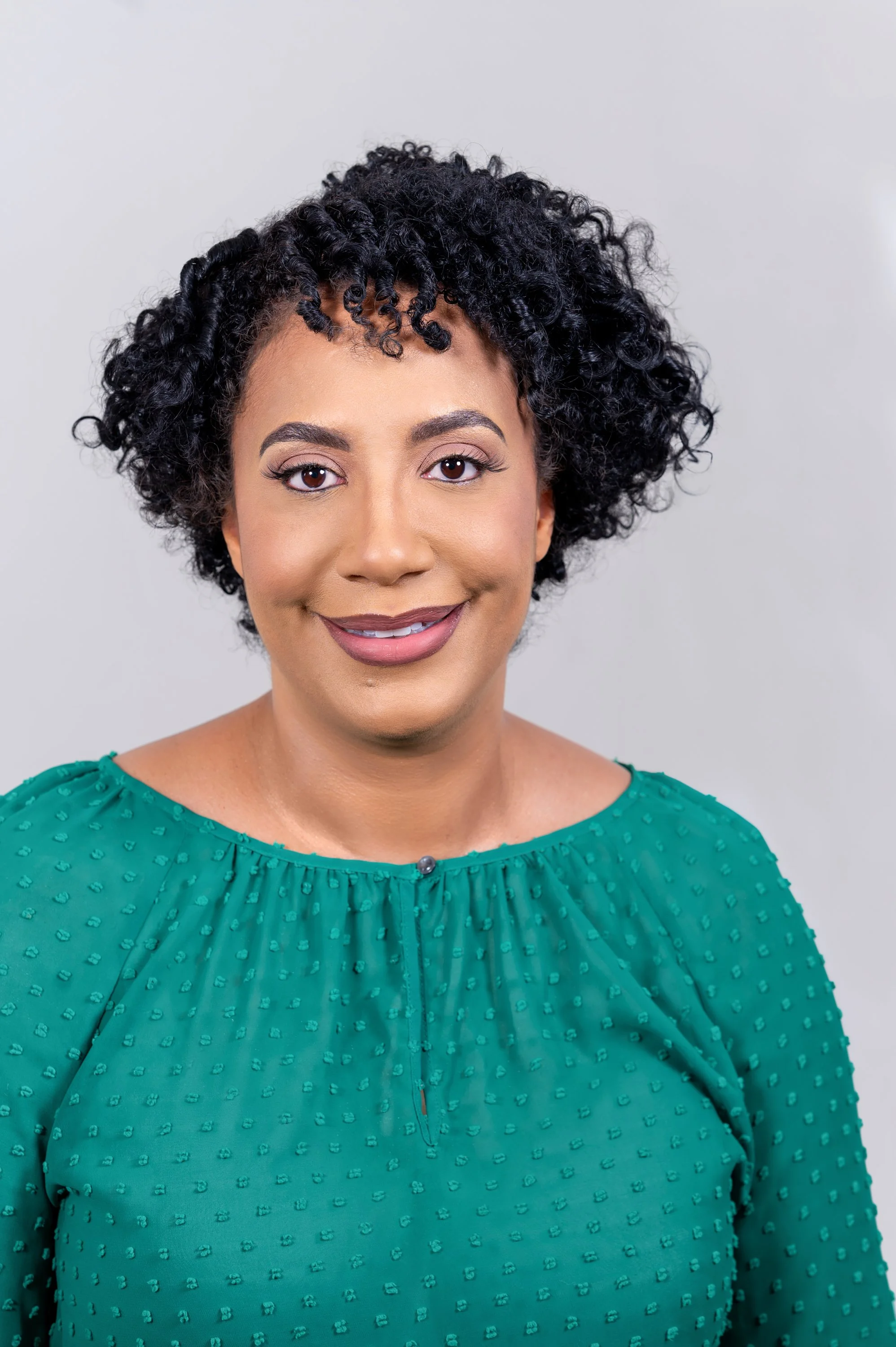 Person with curly hair smiling, wearing a teal textured blouse against a plain background.