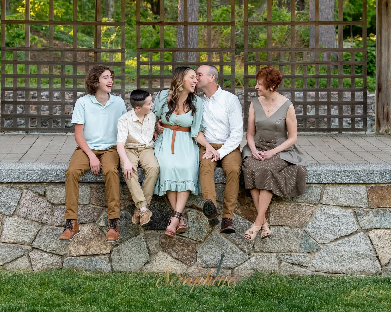 A family of five sitting on a stone ledge in a garden setting, laughing and smiling at each other, with trees and a trellis in the background.