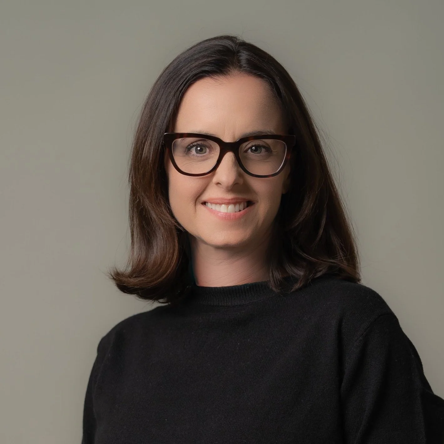 A woman with dark brown hair, wearing black glasses and a black shirt, smiling against a plain gray wall.