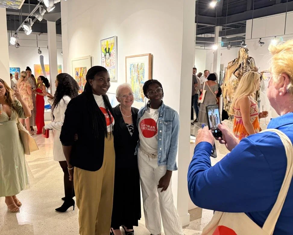 People taking a photo at an art gallery, with several colorful paintings displayed on the white walls behind them.