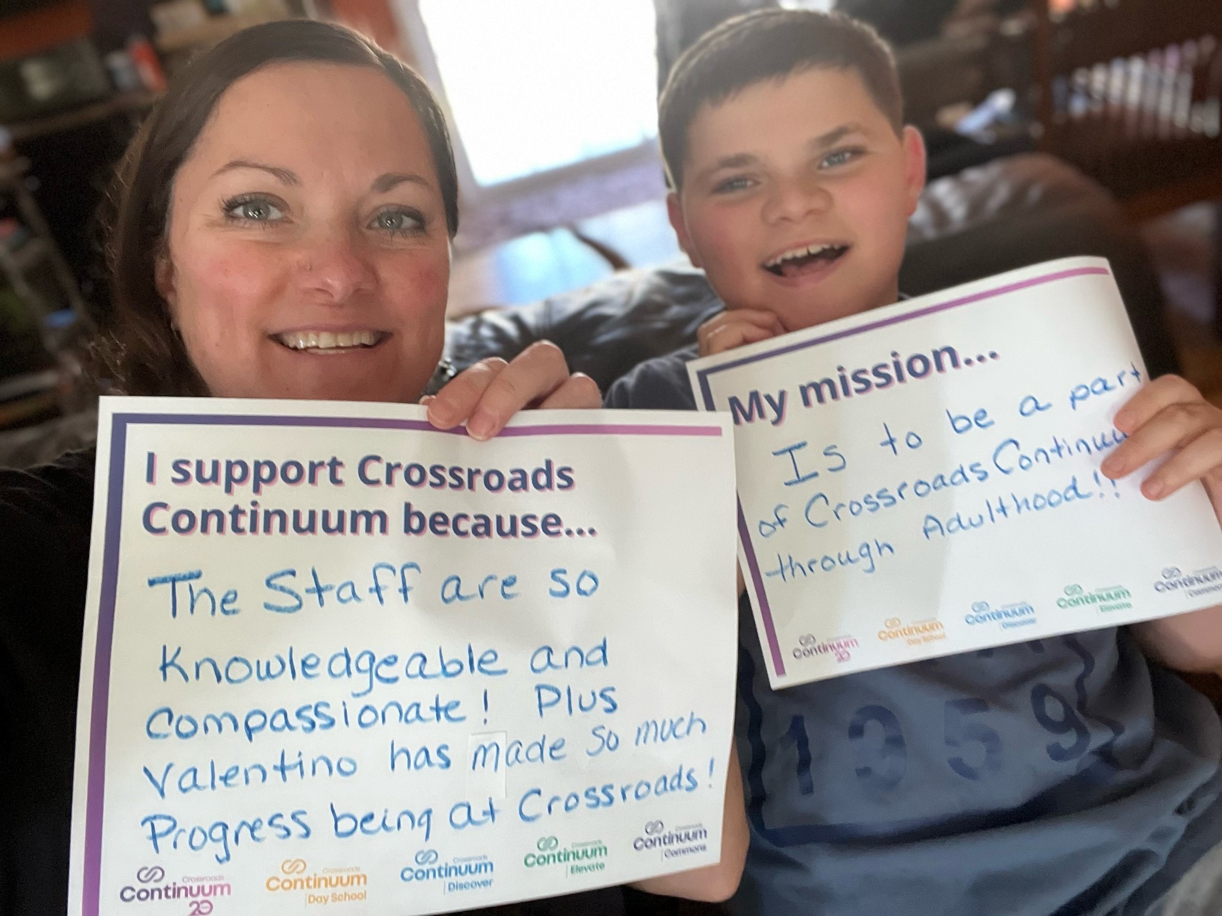 A woman and a boy smiling and holding signs. The woman's sign reads, "I support Crossroads Continuum because... The Staff are so knowledgeable and compassionate! Plus Valentino has made so much progress being at Crossroads!" The boy's sign reads, "My