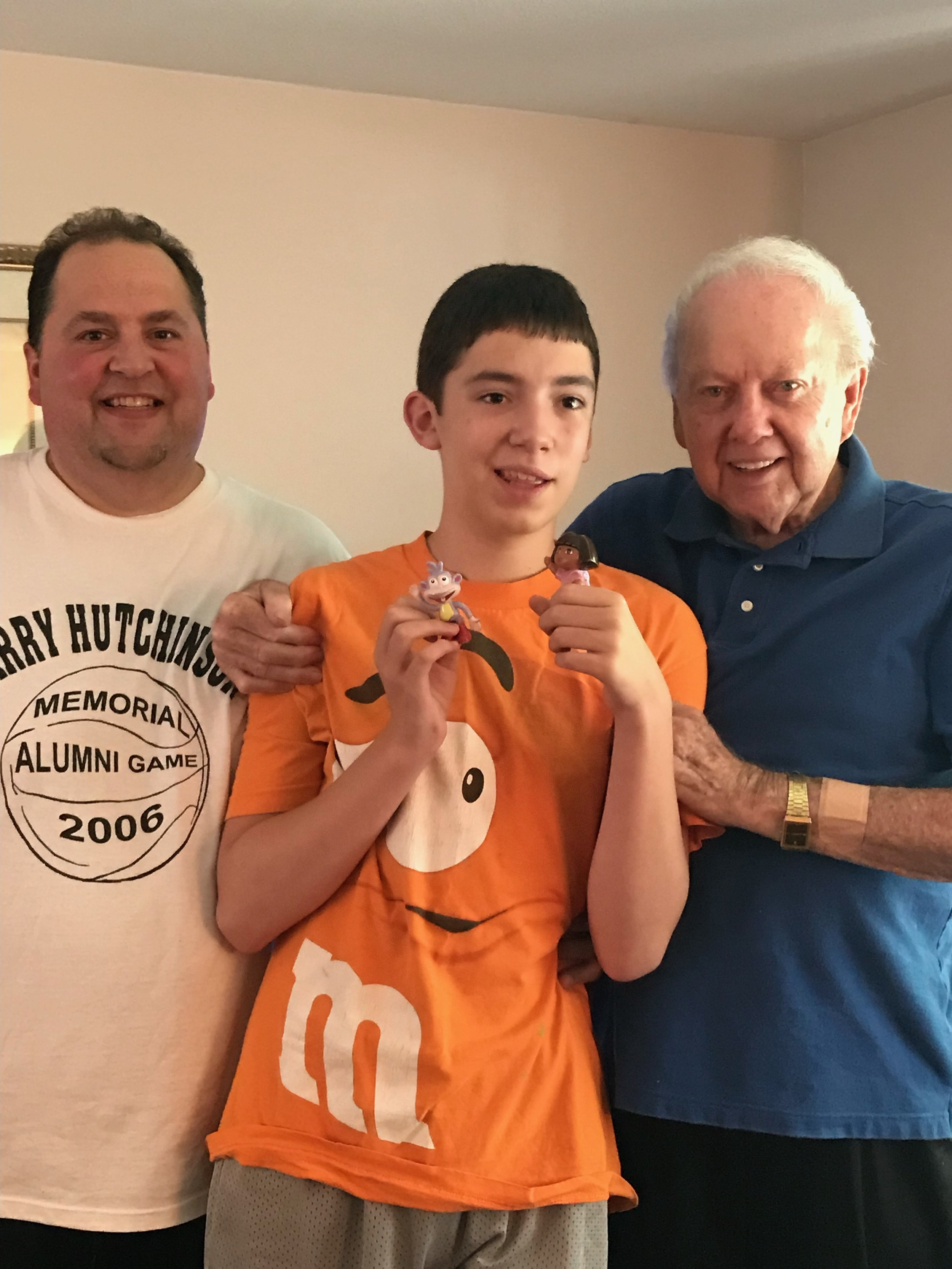 Three people posing together indoors, with the person in the center holding small cartoon figurines. The left person is wearing a white t-shirt, the center person is wearing an orange t-shirt, and the right person is wearing a blue shirt. They are al