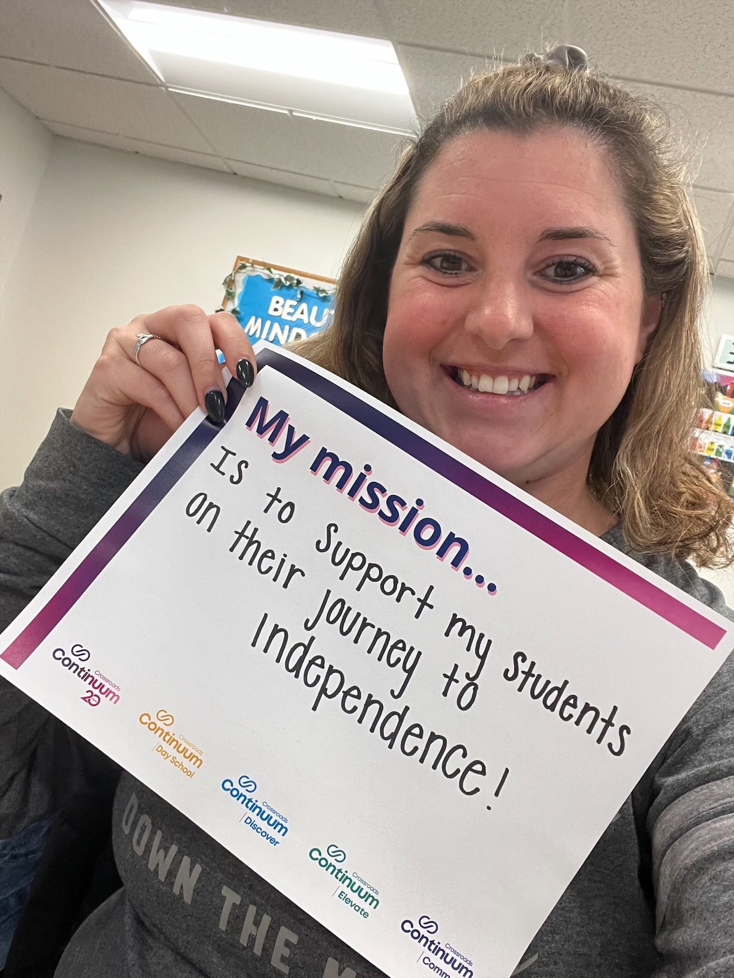 A person holding a sign that reads, "My mission... Is to support my students on their journey to independence!" The sign includes logos for Continuum Day School, Continuum Discover, Continuum Elevate, and Continuum Community. The individual is smilin