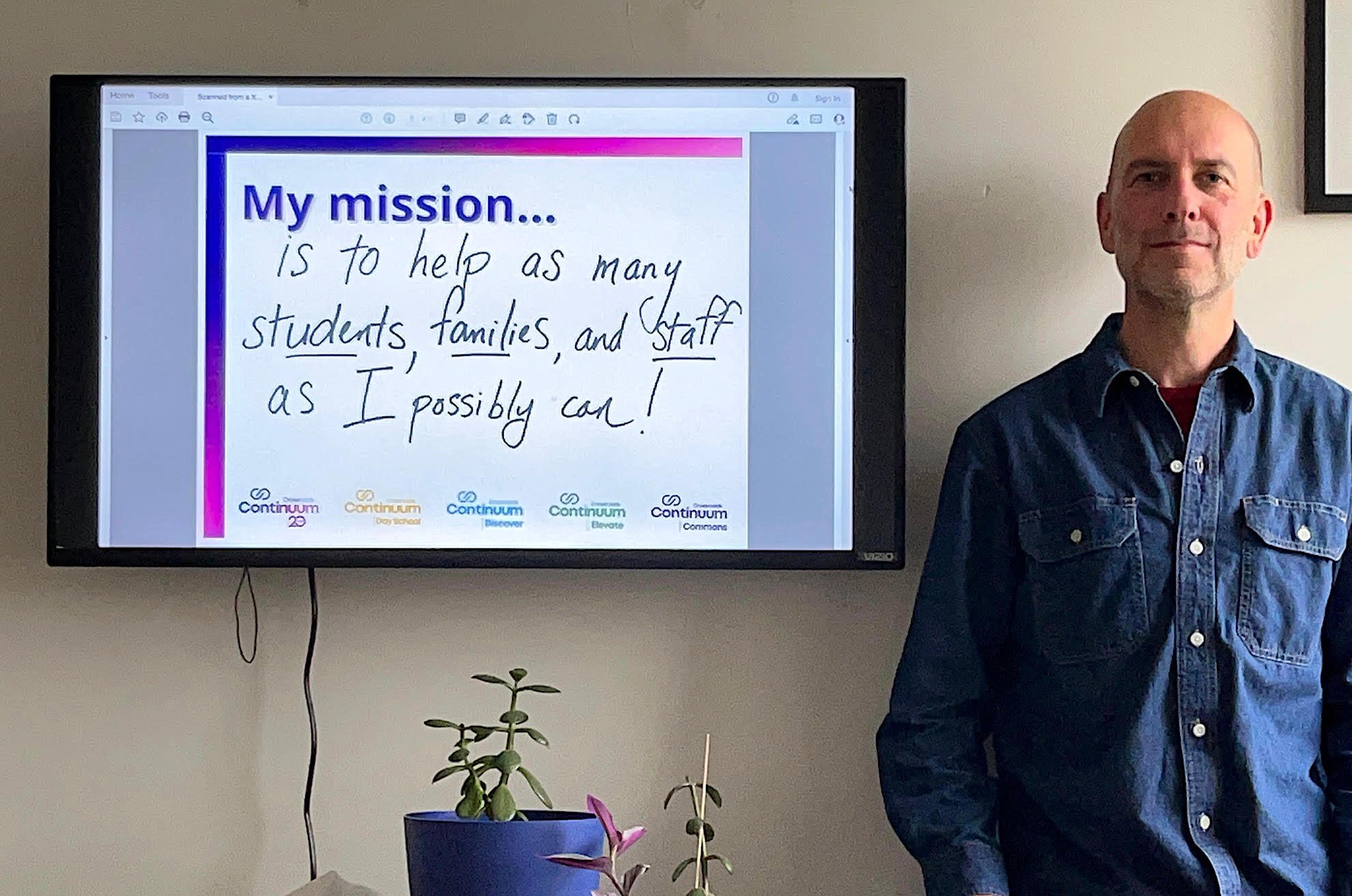 A man standing next to a screen displaying a slide with the text: 'My mission is to help as many students, families, and staff as I possibly can!' Below the text, there are logos for Continuum 2.0, Continuum Day School, Continuum Discover, Continuum