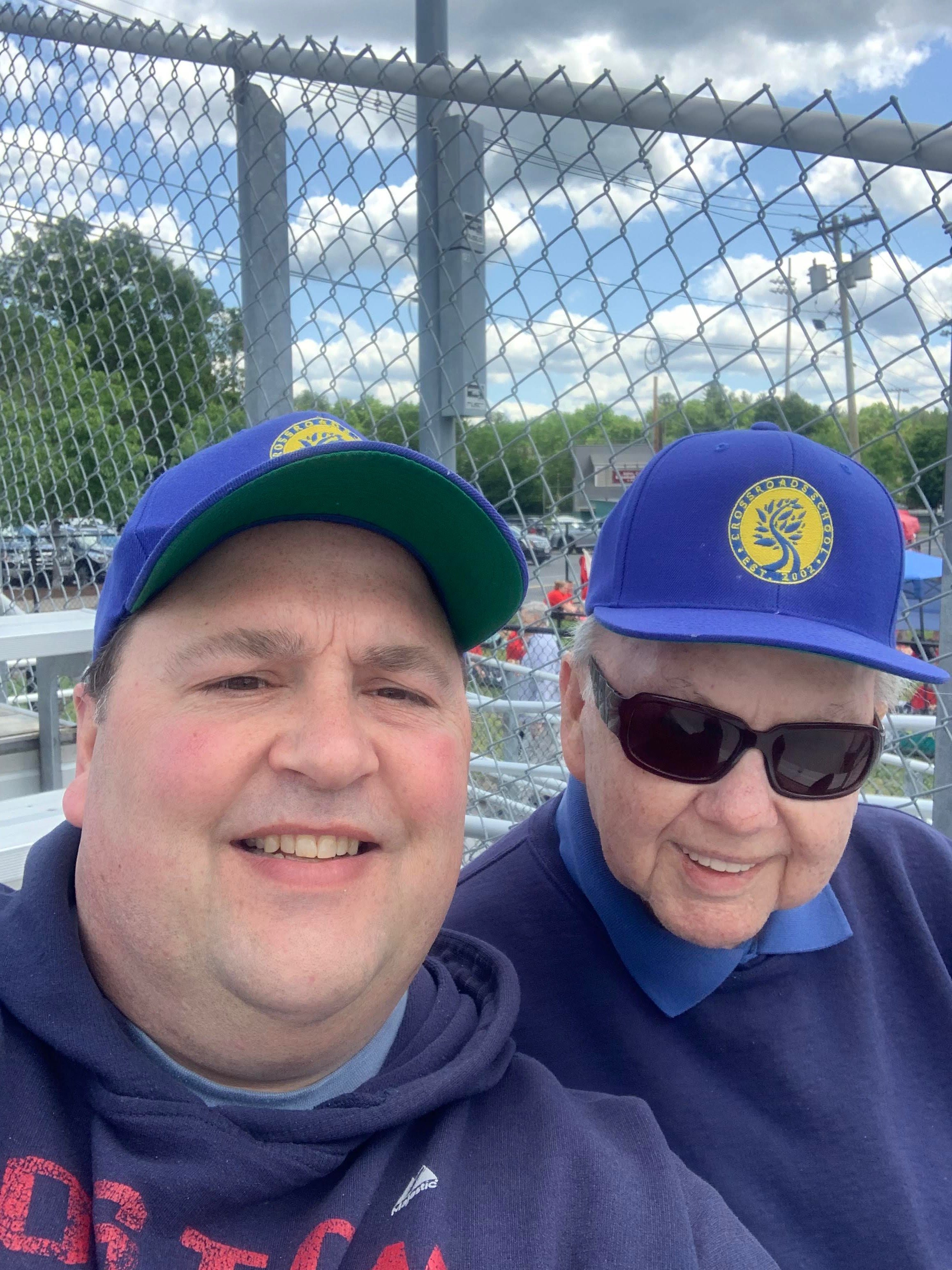 Two people wearing blue baseball caps sitting in front of a chain-link fence at an outdoor event.