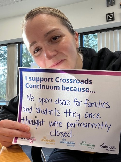 Person holding a sign that reads "I support Crossroads Continuum because... We open doors for families and students they once thought were permanently closed." Background shows windows with trees outside.