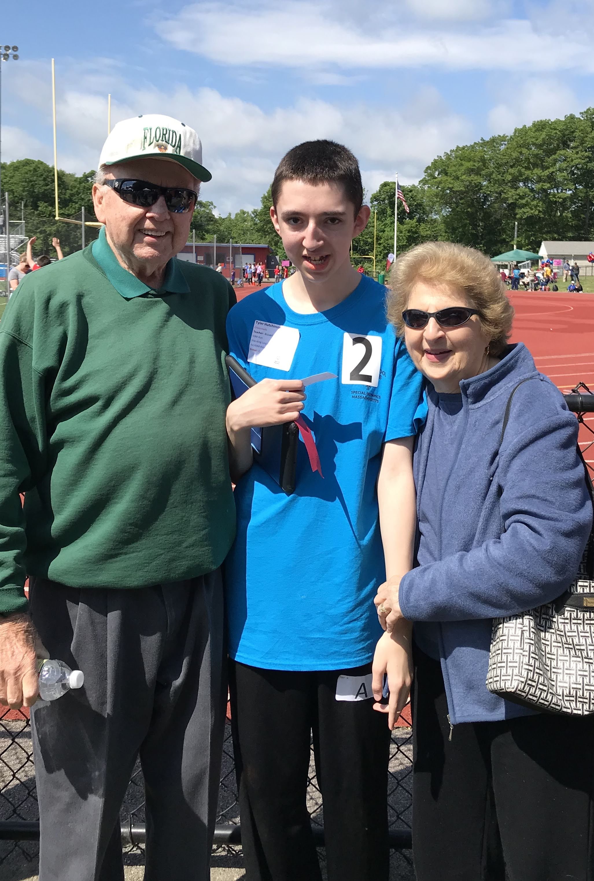 Three people at a track and field event: a person in a blue shirt with the number 2 stands between two older adults, one wearing a green sweater and hat, the other wearing a blue jacket. They are outdoors with a running track and spectators in the ba