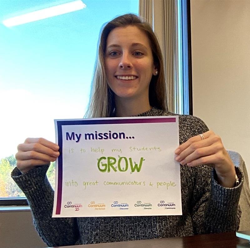 Person smiling while holding a sign that reads, "My mission is to help my students GROW into great communicators & people."