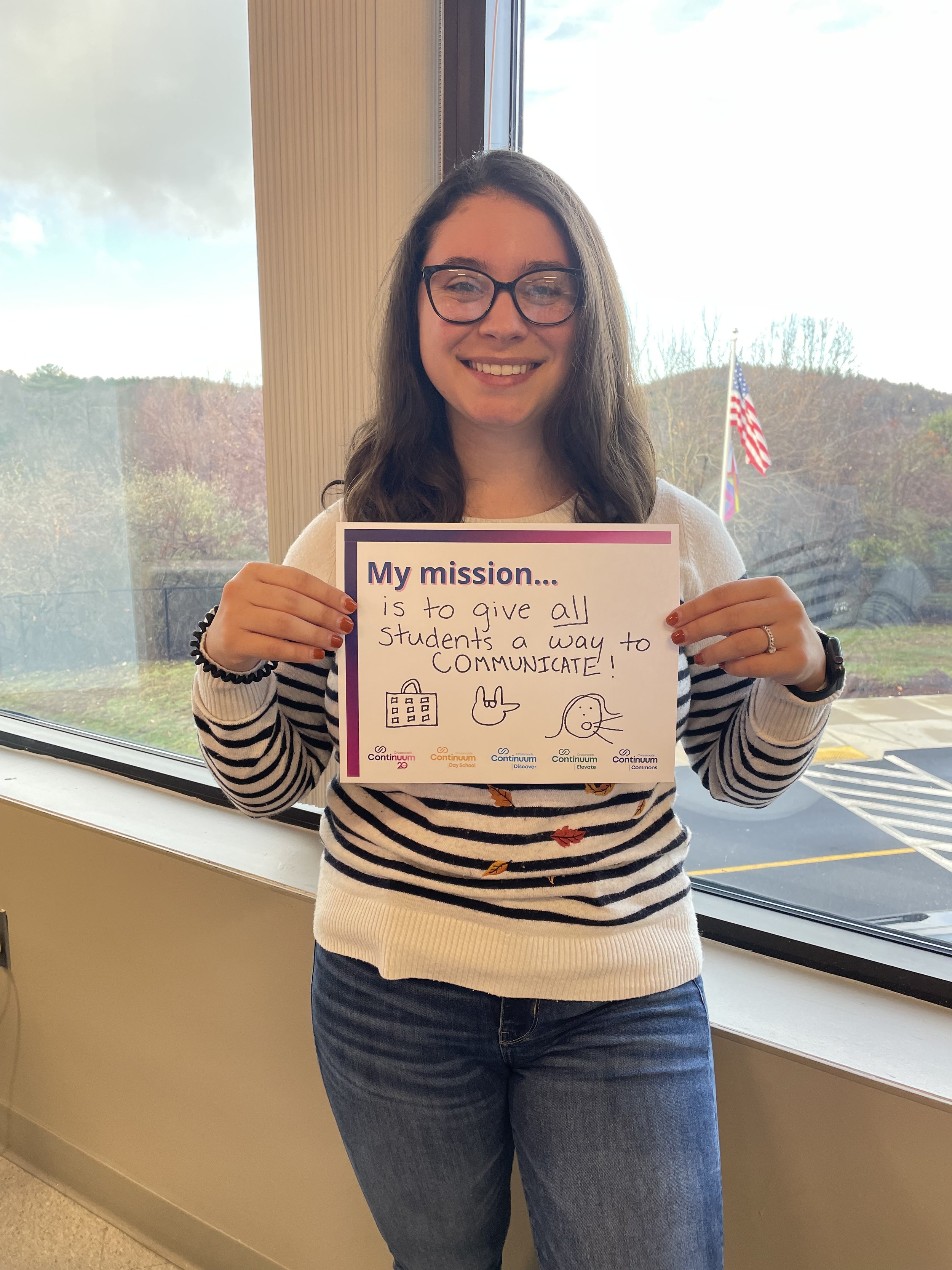 A person wearing glasses and a striped sweater is standing indoors by a window, holding a sign that reads, "My mission is to give all students a way to communicate!" The background shows an American flag and trees outside.