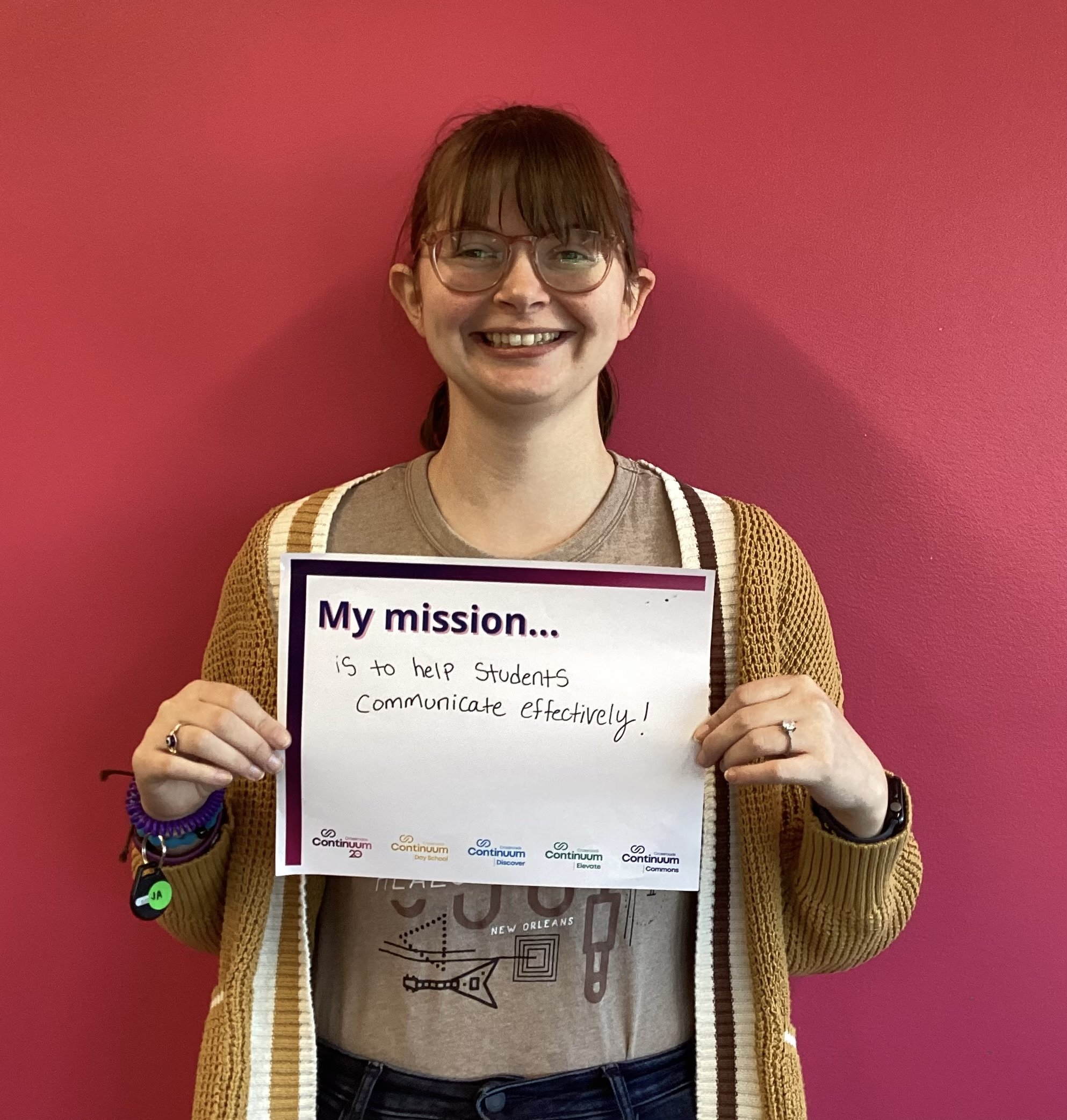 Person holding a sign that reads, 'My mission... is to help students communicate effectively!' standing in front of a pink wall.