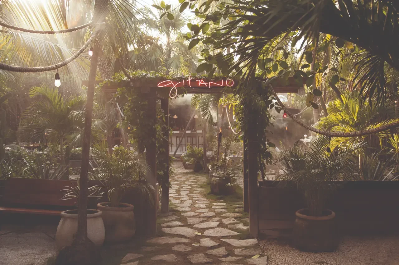 Sunlight filters through lush jungle foliage at the original entrance of GITANO JUNGLE in Tulum, featuring a stone path, tropical plants, and the iconic pink neon ‘GITANO’ sign hanging above a rustic wooden pergola.