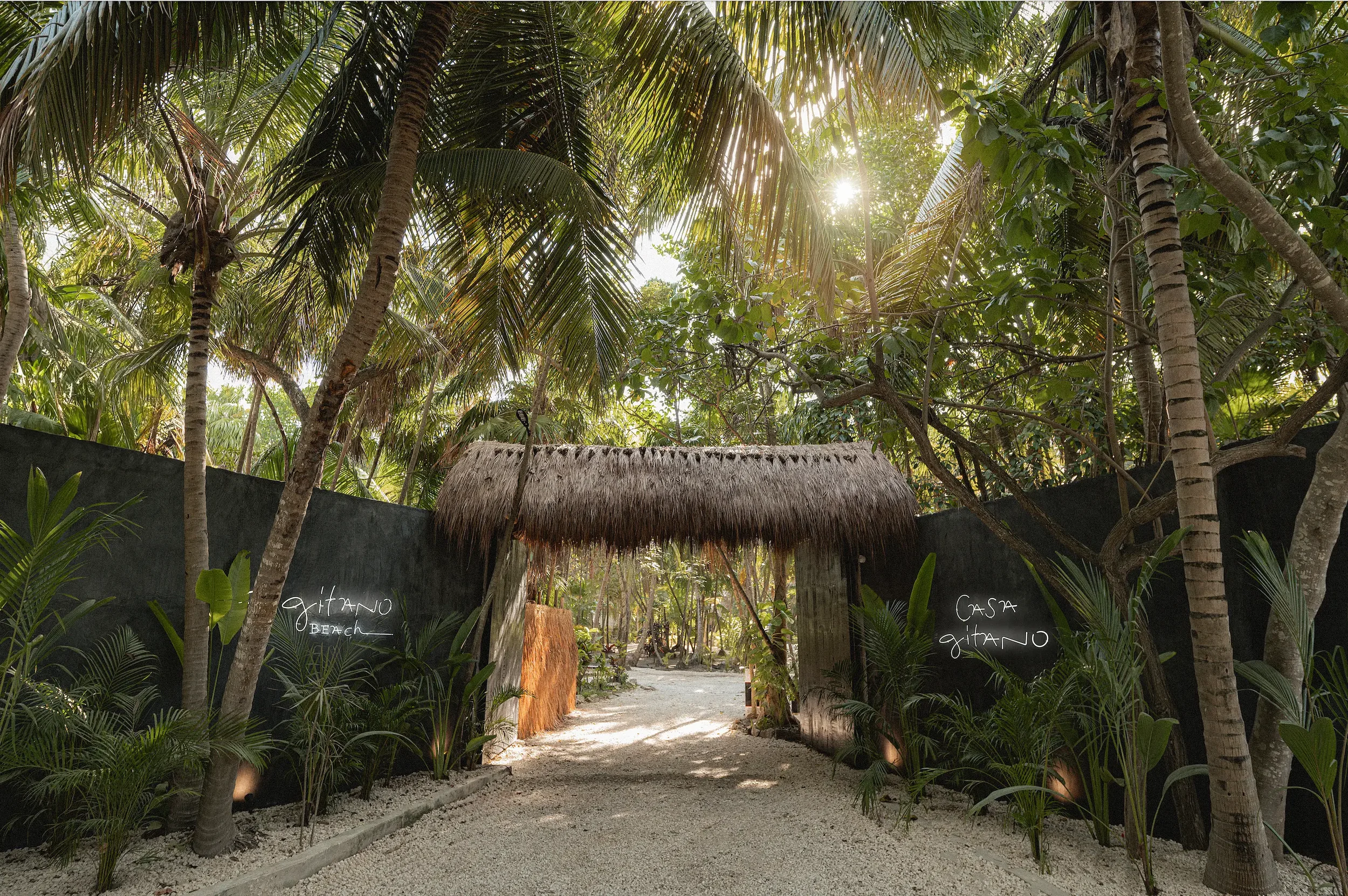 Entrance of CASA GITANO and GITANO BEACH, surrounded by palms and lush greenery and two white neon signs.