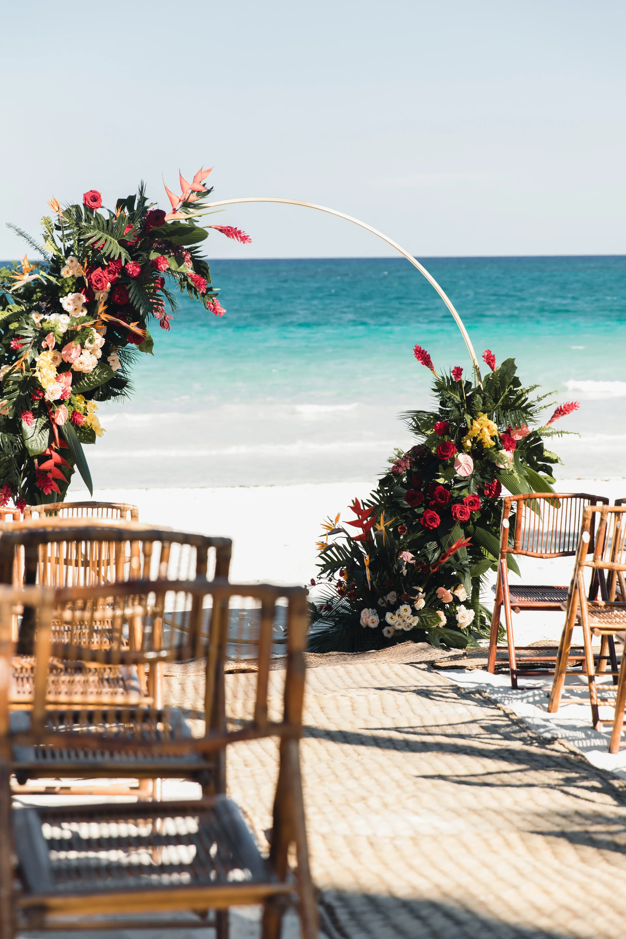 Elegant beachfront wedding setup at CASA GITANO, Tulum — a tropical floral arch with vibrant red, pink, and white blooms frames the turquoise Caribbean Sea, with wooden chairs arranged on pristine white sand