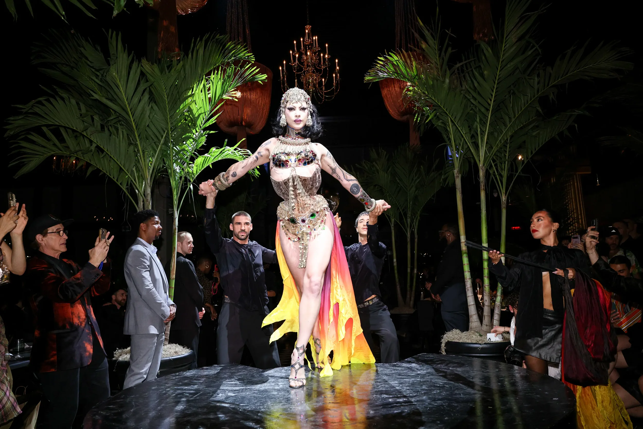 Drag performer in a jeweled costume takes center stage at GITANO NYC, surrounded by dancers and guests under dramatic chandeliers and lush tropical palms during a vibrant nightlife event.