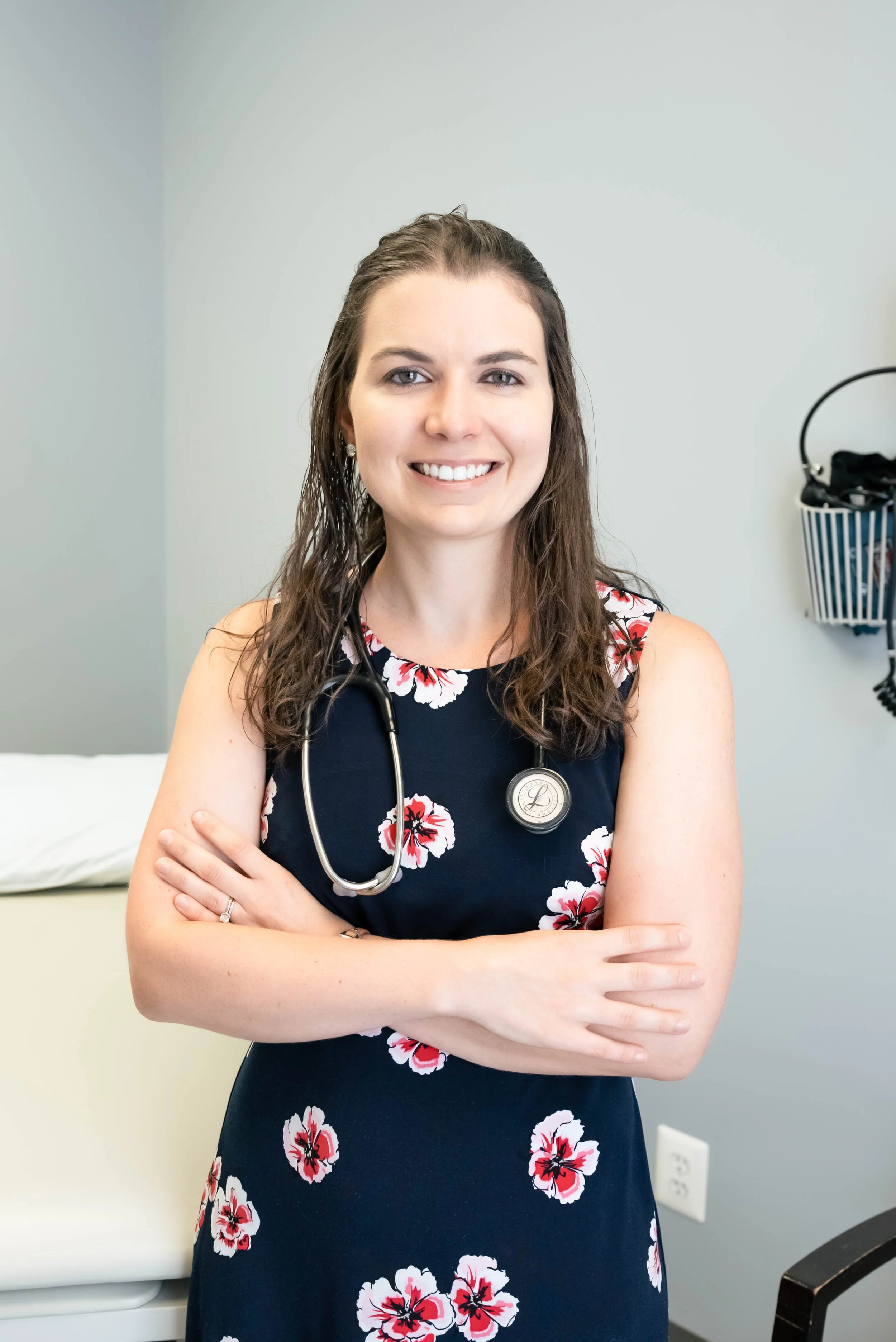 Female healthcare professional with stethoscope, smiling, in a floral dress, standing in a medical office.