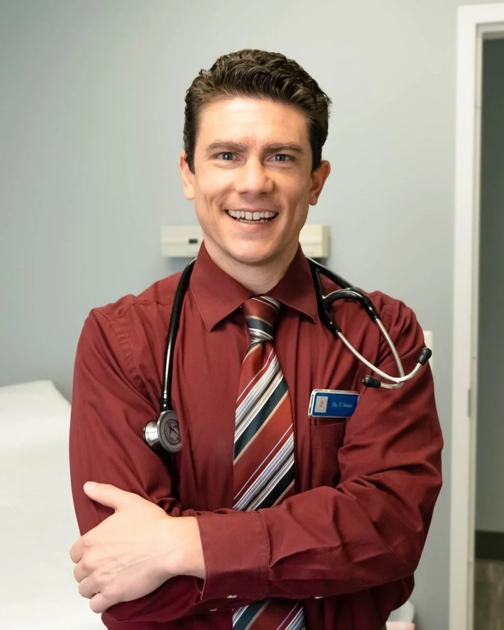 A male doctor smiling with a stethoscope around his neck, wearing a burgundy shirt and striped tie in a medical office.