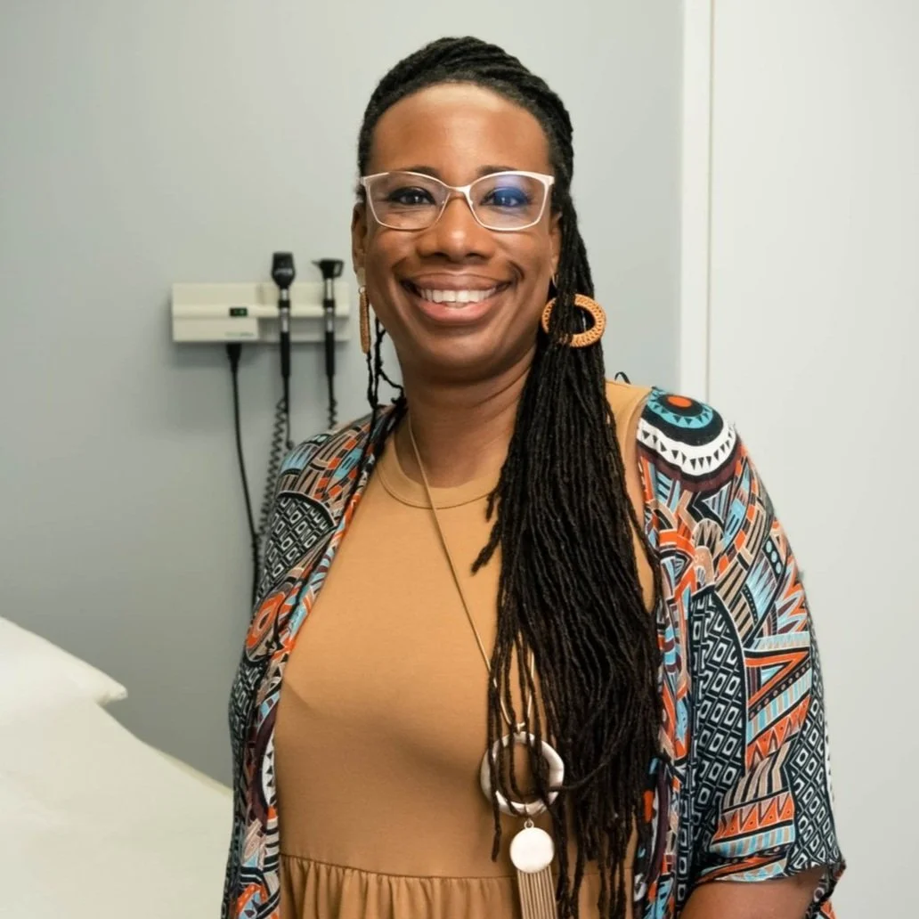 A woman with long dreadlocks and glasses smiling in a medical office, wearing a patterned cardigan and brown dress.