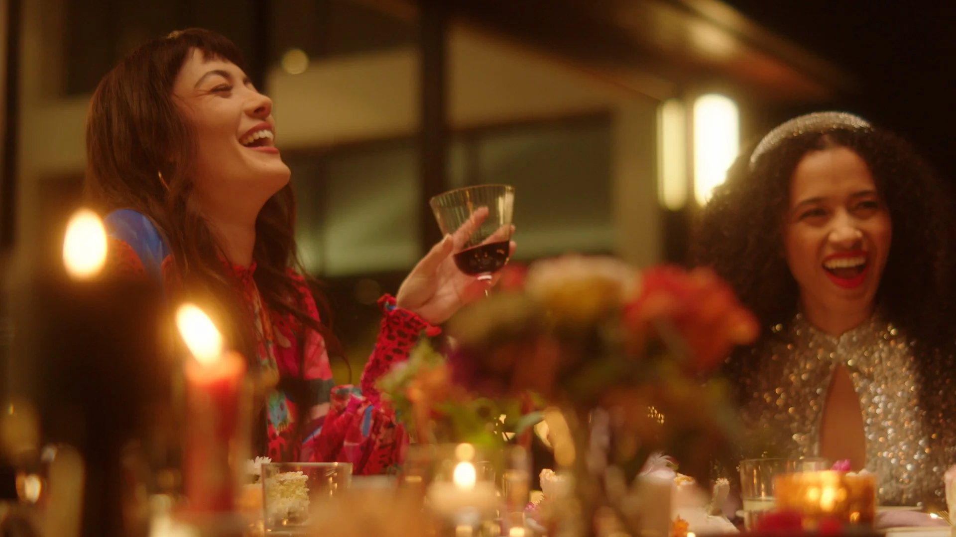 Two women laugh and enjoy a celebration at a dinner table, with candles and flowers, one holding a glass of red wine.